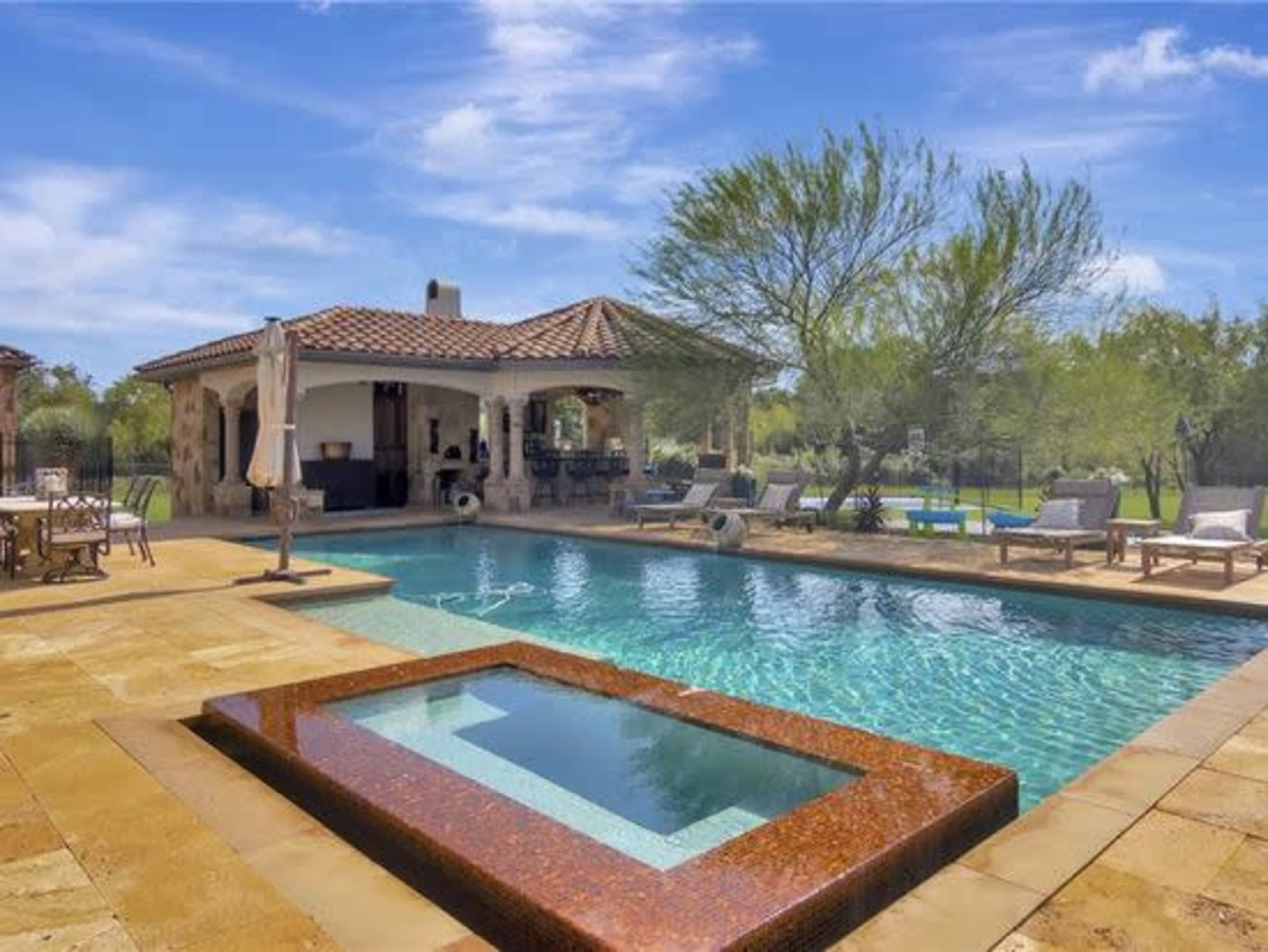 A spacious outdoor pool area featuring a rectangular pool with a spa, surrounded by lounge chairs and a covered patio with dining space.