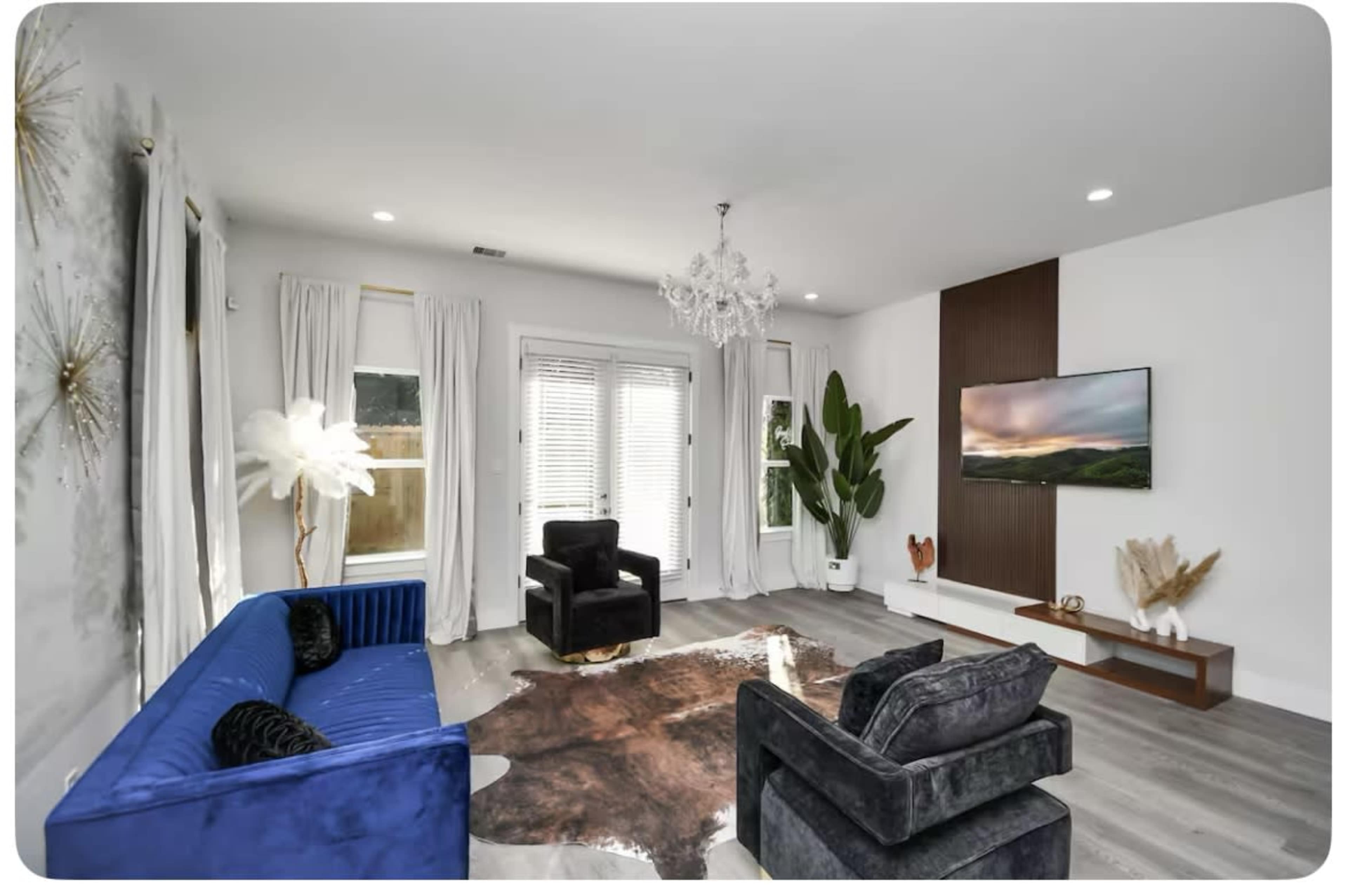 The living room features a blue sofa, two black chairs, a cowhide rug, a large plant, and a wall-mounted television.