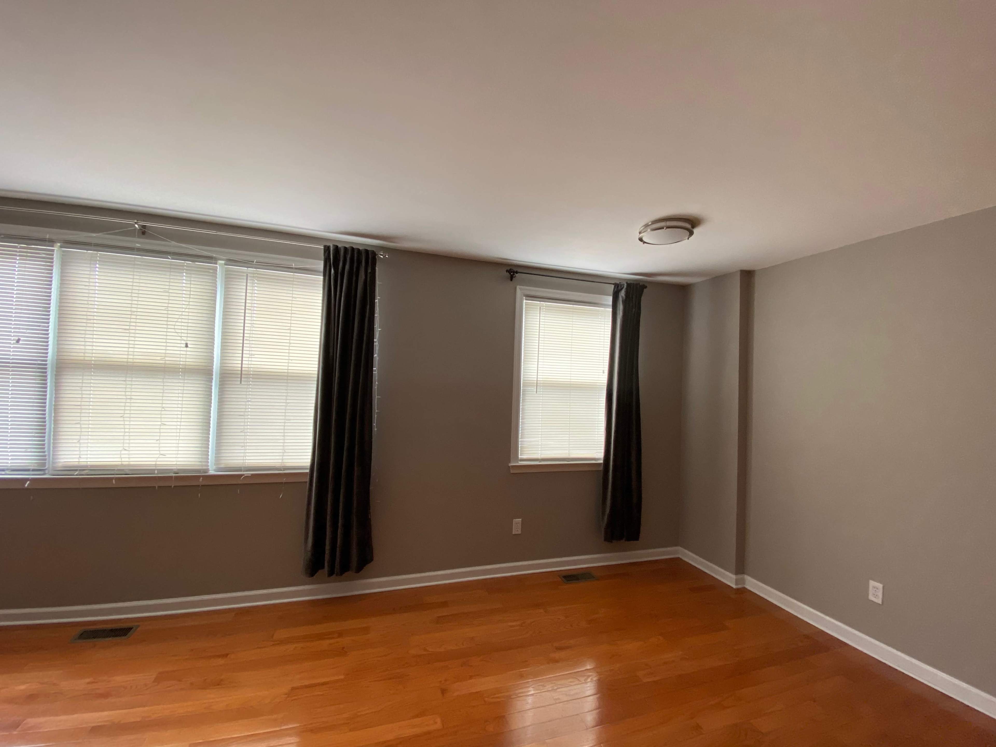 An empty room with hardwood flooring, two windows with blinds, and dark curtains.