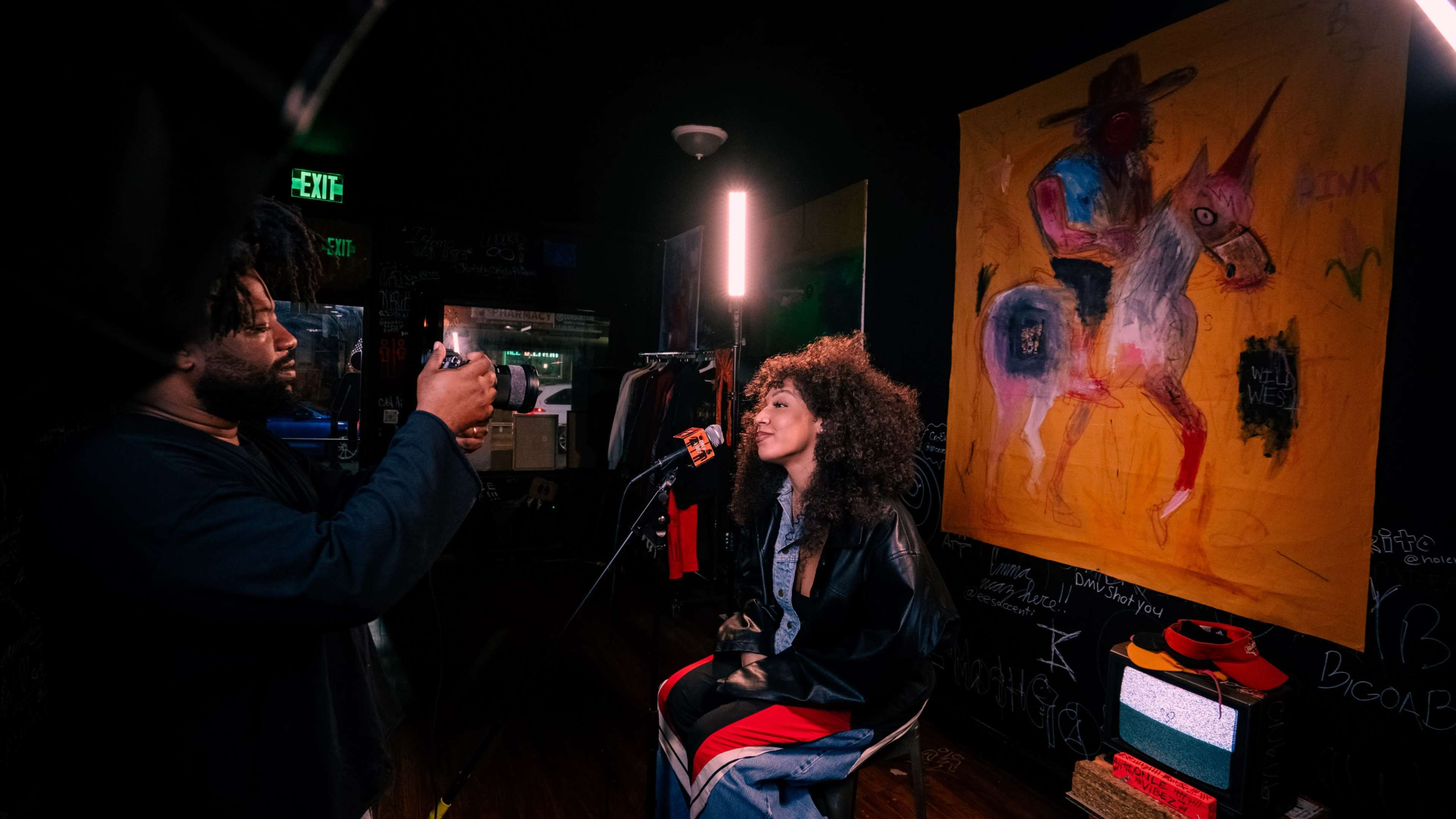 A person is filming a woman seated in front of a colorful mural inside a dimly lit room.