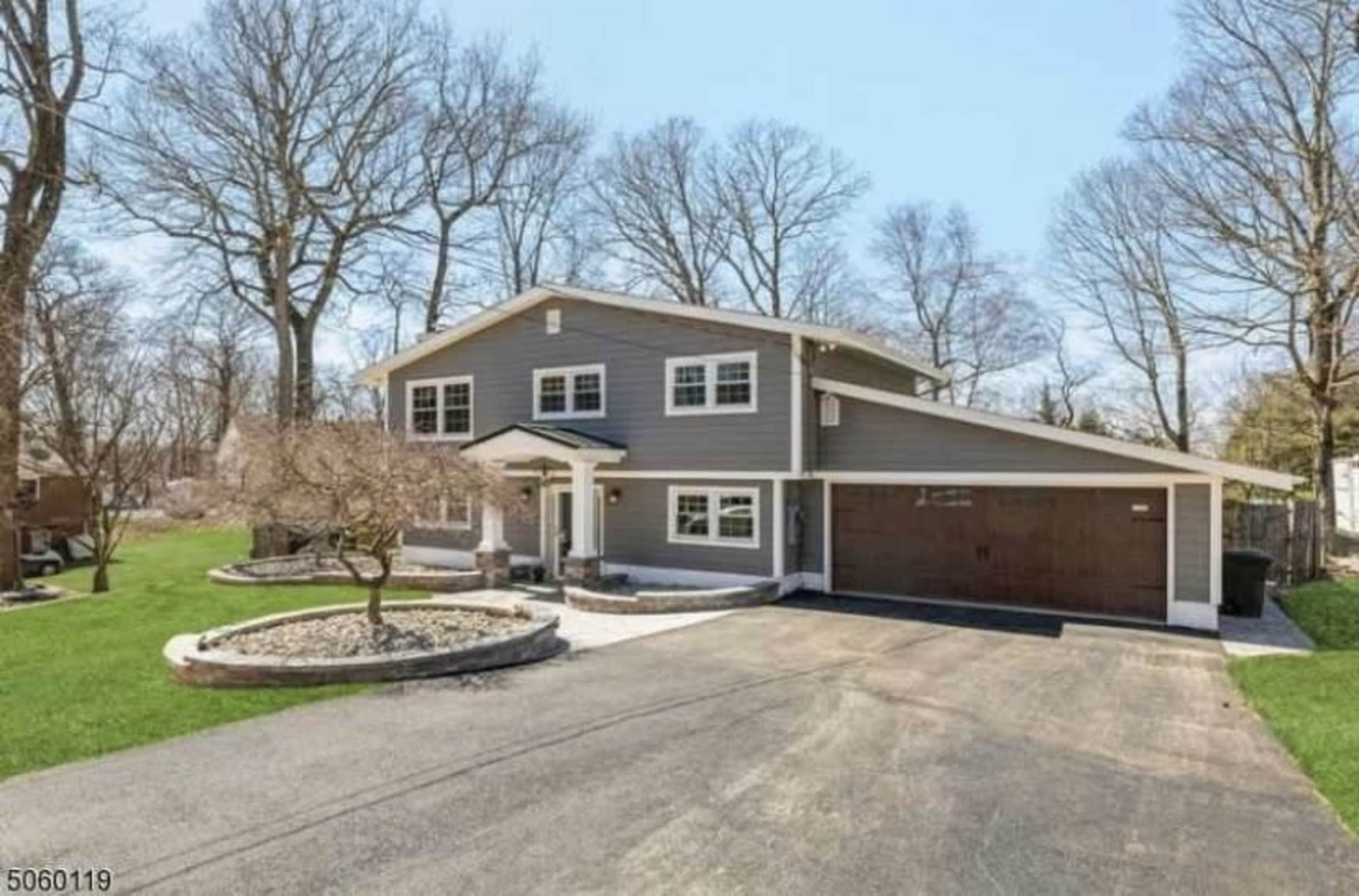 The image shows a two-story gray house with a garage, situated on a landscaped property with trees and a clear blue sky.