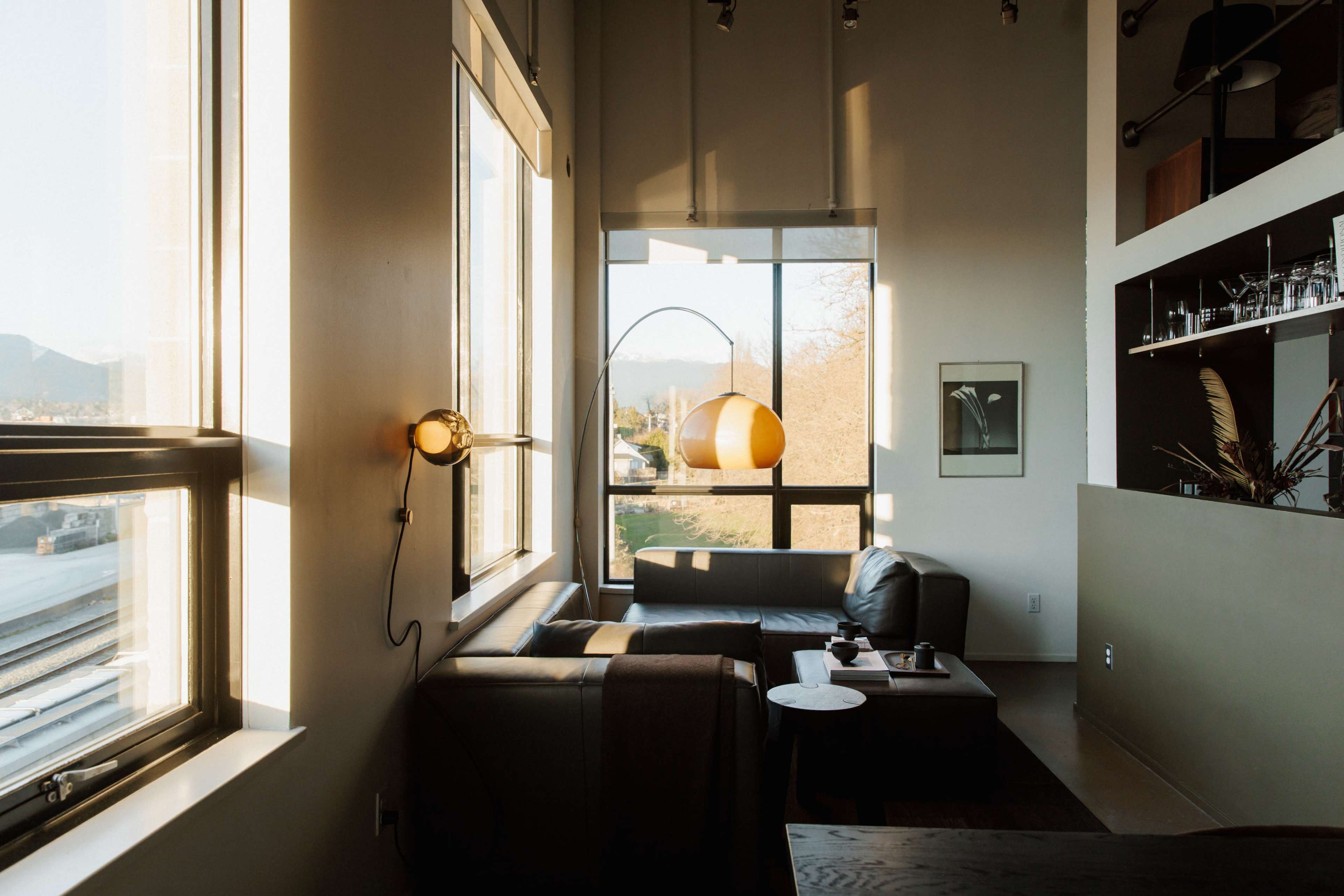 The image shows a modern living room with a dark leather sofa, a coffee table, and large windows allowing natural light to fill the space.