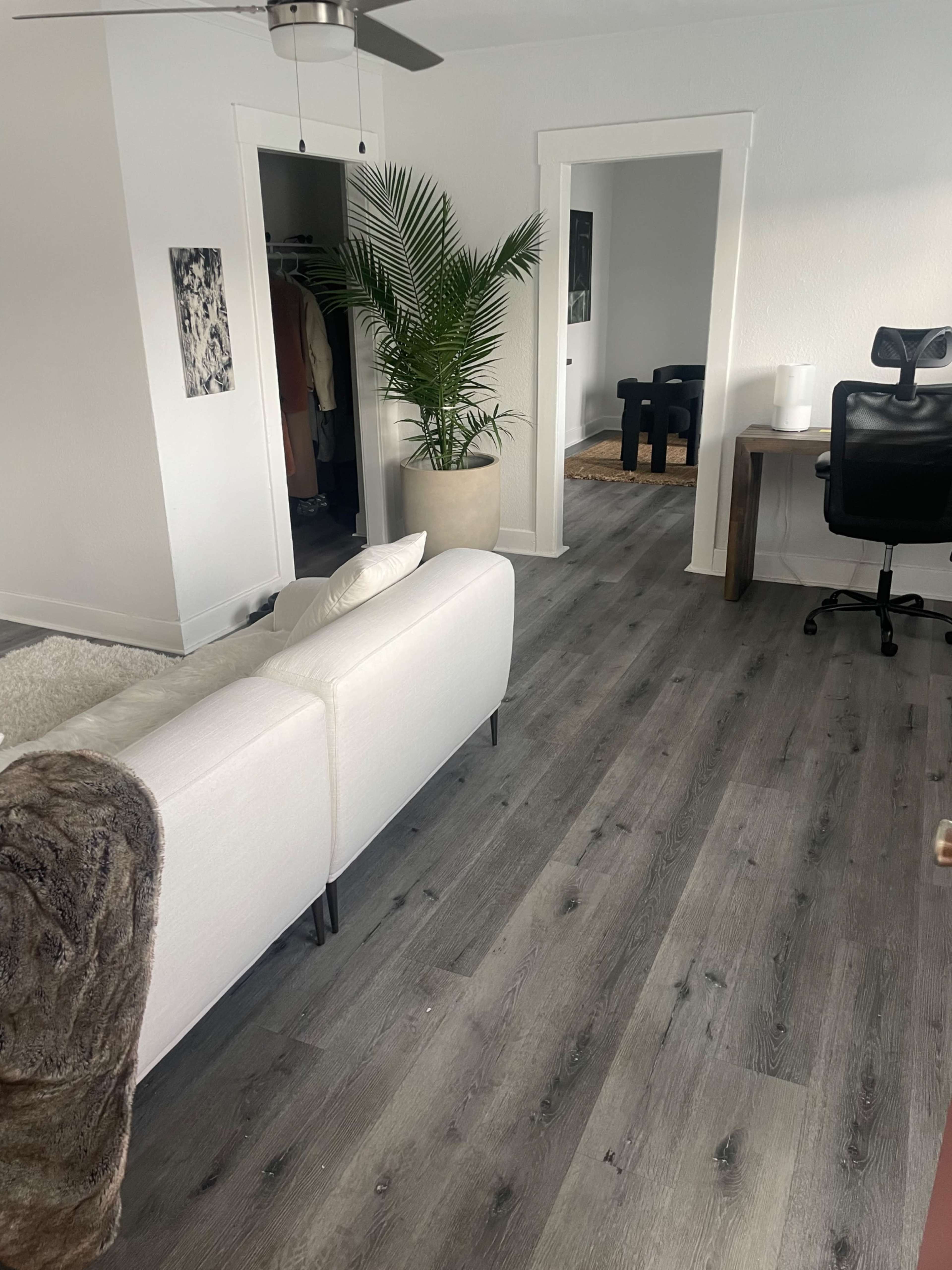 The image shows a bright living space with a white sofa, a large potted plant, and a wooden desk with a chair, along with a view of an adjacent room.