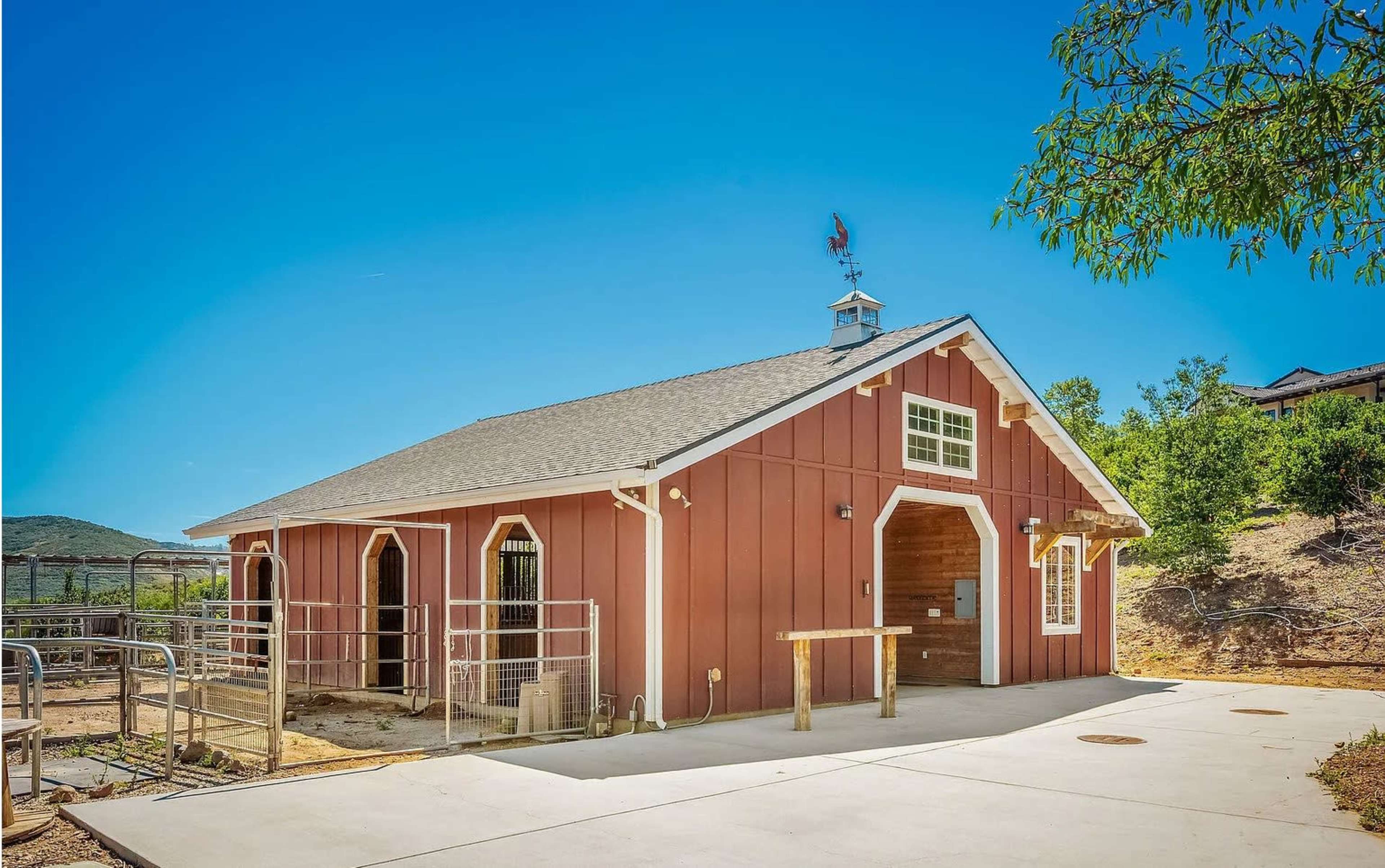 The image shows a red barn-style building with white trim, surrounded by a dirt area and trees under a clear blue sky.