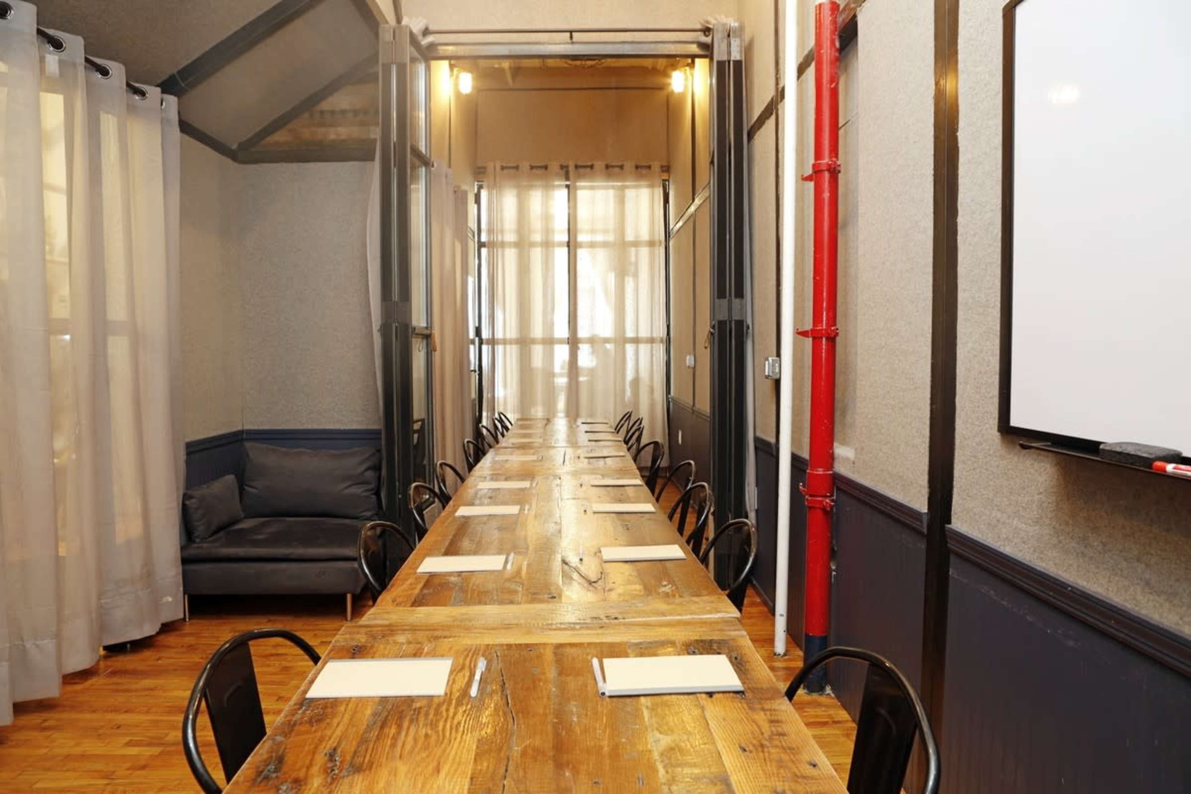 The image shows a long wooden table set for a meeting, flanked by black chairs, with large windows covered by sheer curtains allowing light to enter the room.