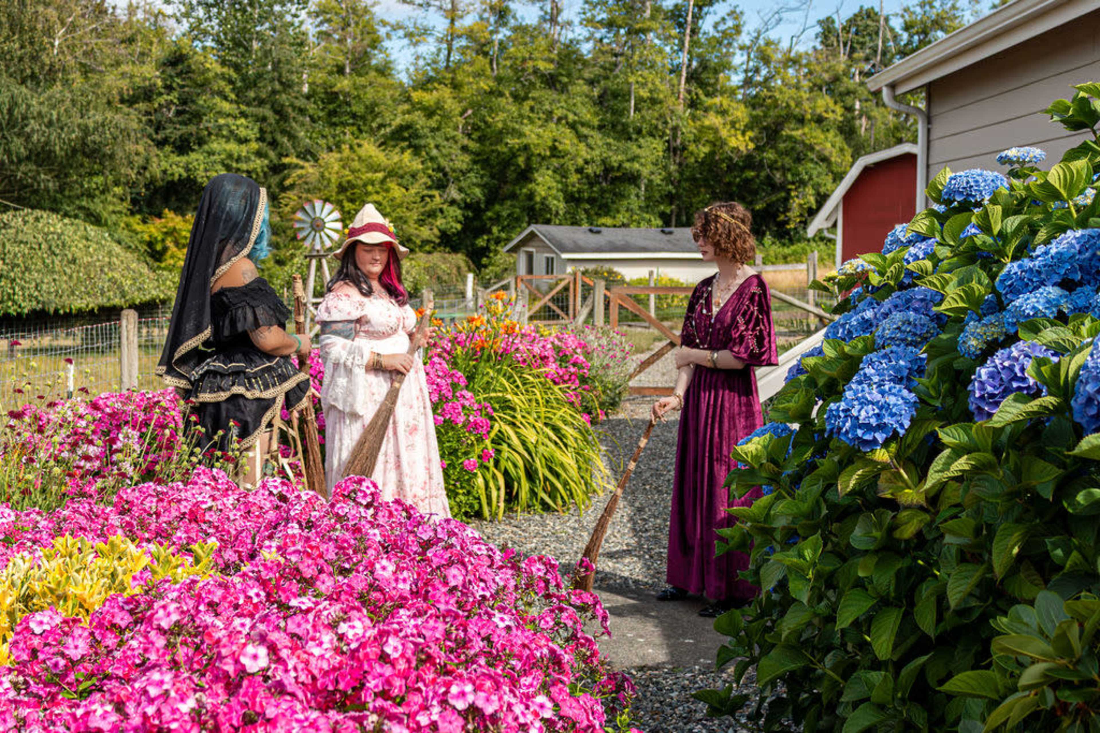 Three women dressed in colorful clothing stand in a flower garden filled with vibrant pink and blue blooms, surrounded by greenery and wooden fences.