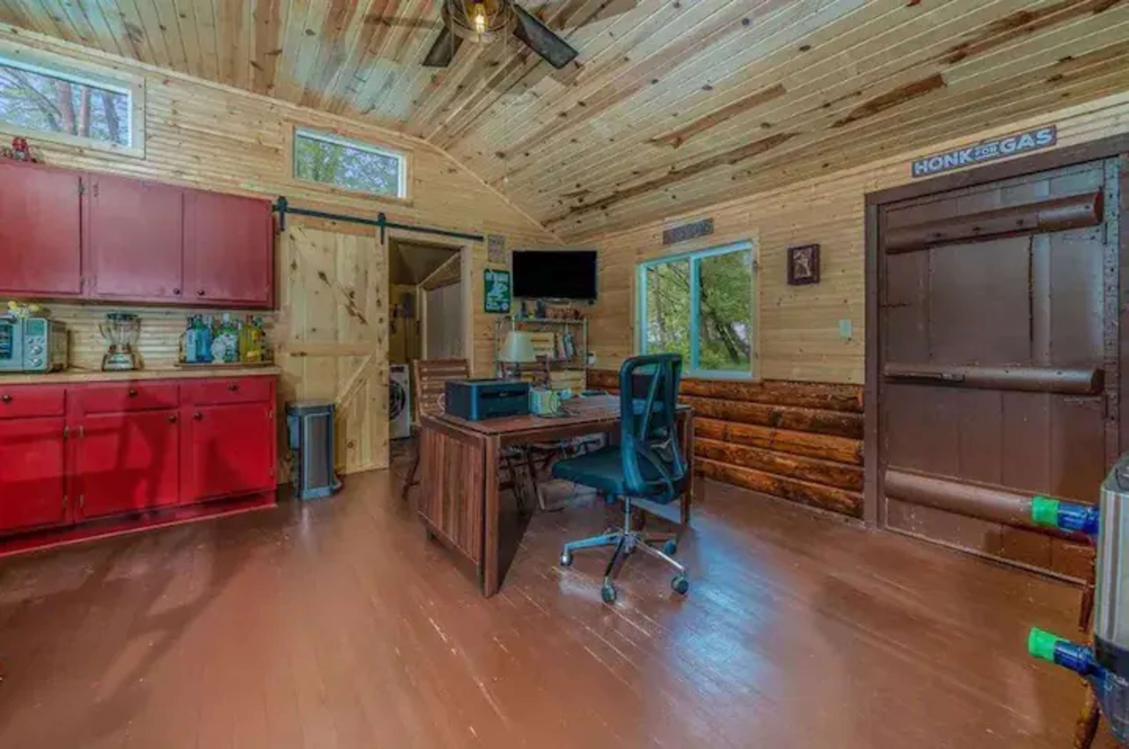 A small wooden cabin interior with a desk, chair, red cabinets, and a door leading outside.