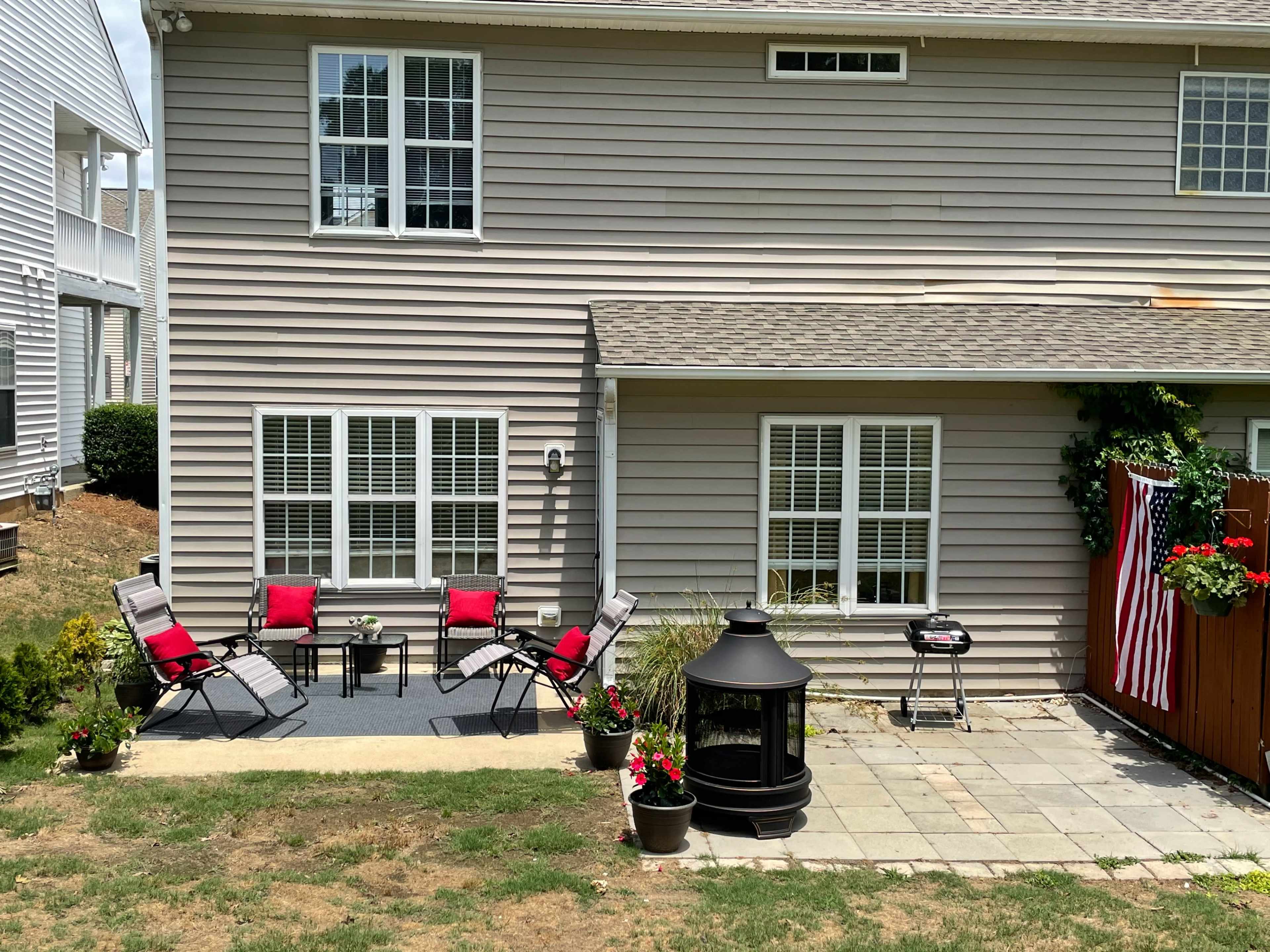 The image shows a patio area behind a home with a set of red-cushioned chairs, a small table, a grill, and planters with flowers.