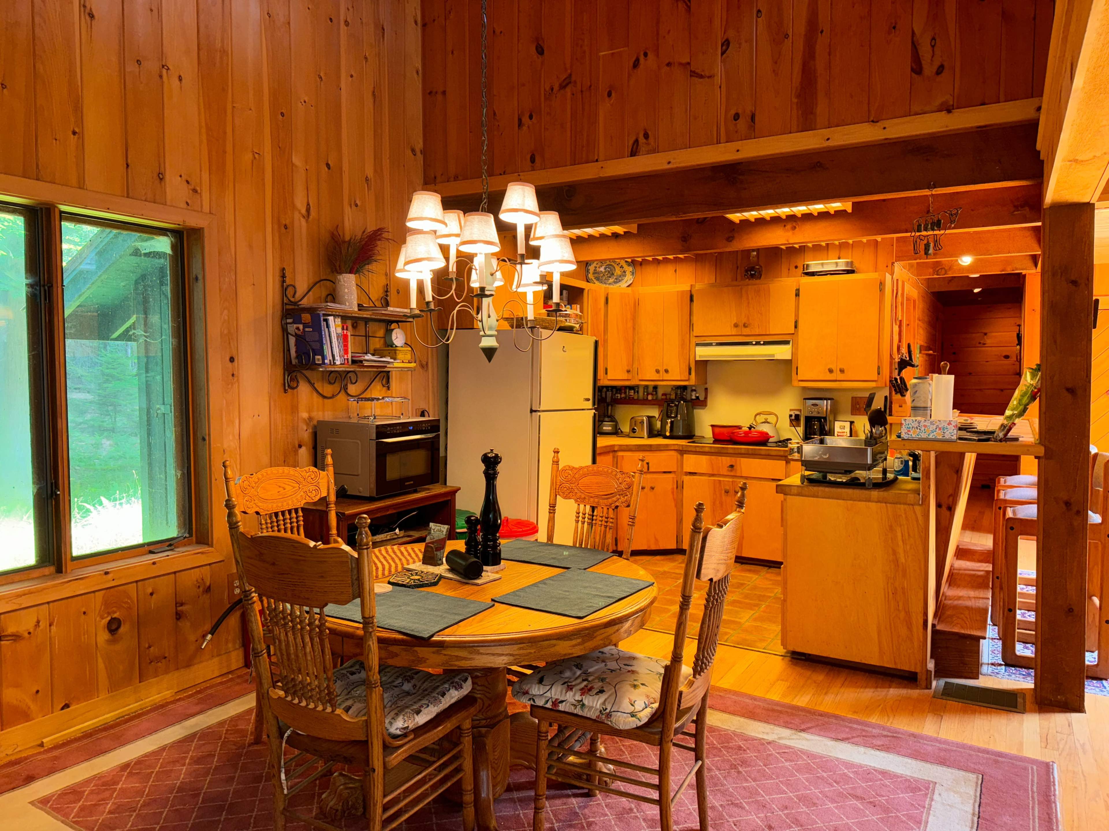 The image shows a rustic kitchen and dining area with wooden walls, a round dining table set for four, and a well-equipped kitchen visible in the background.
