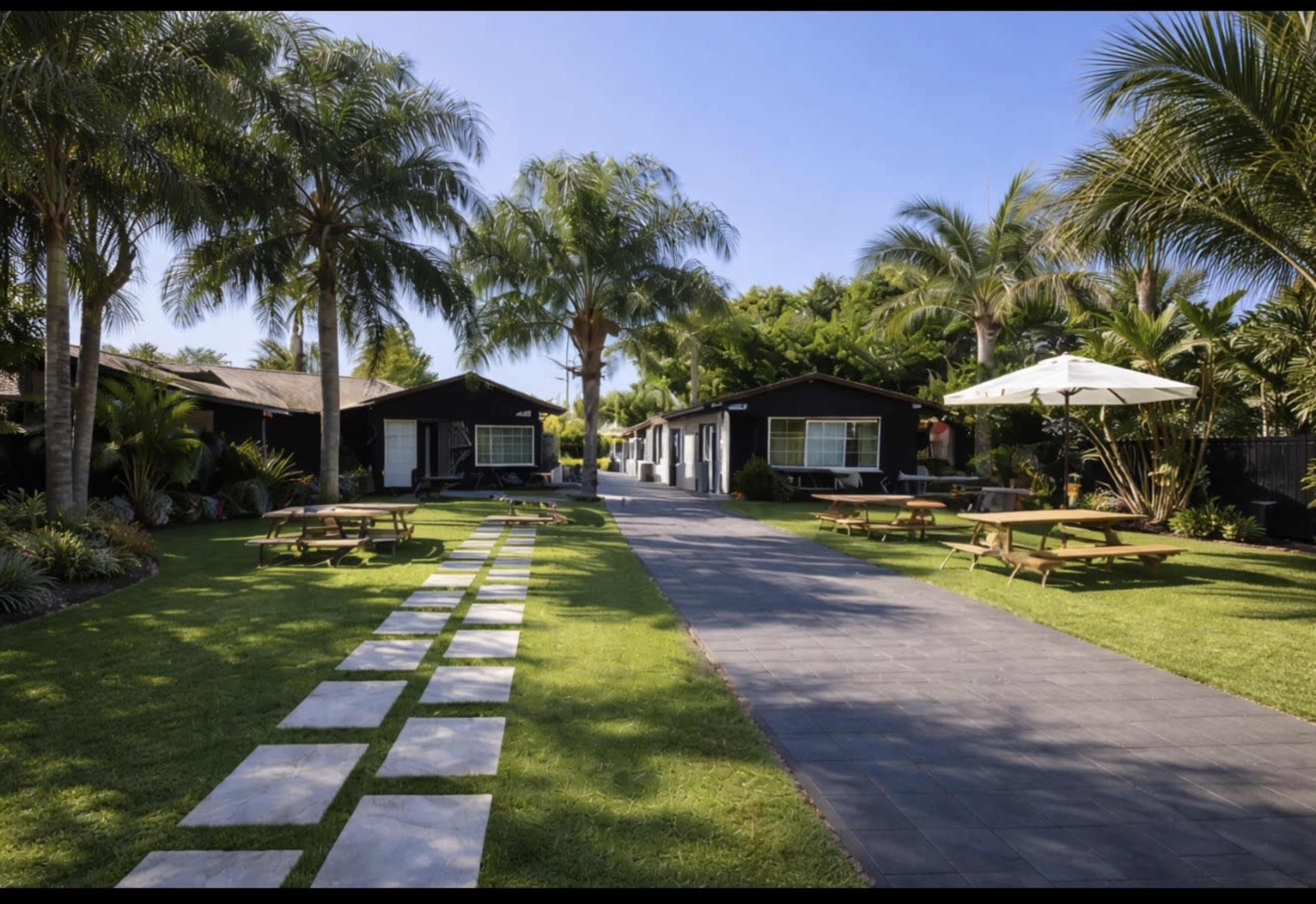 The image shows a well-maintained outdoor area with several small cabins, a pathway of stepping stones, and picnic tables under palm trees.