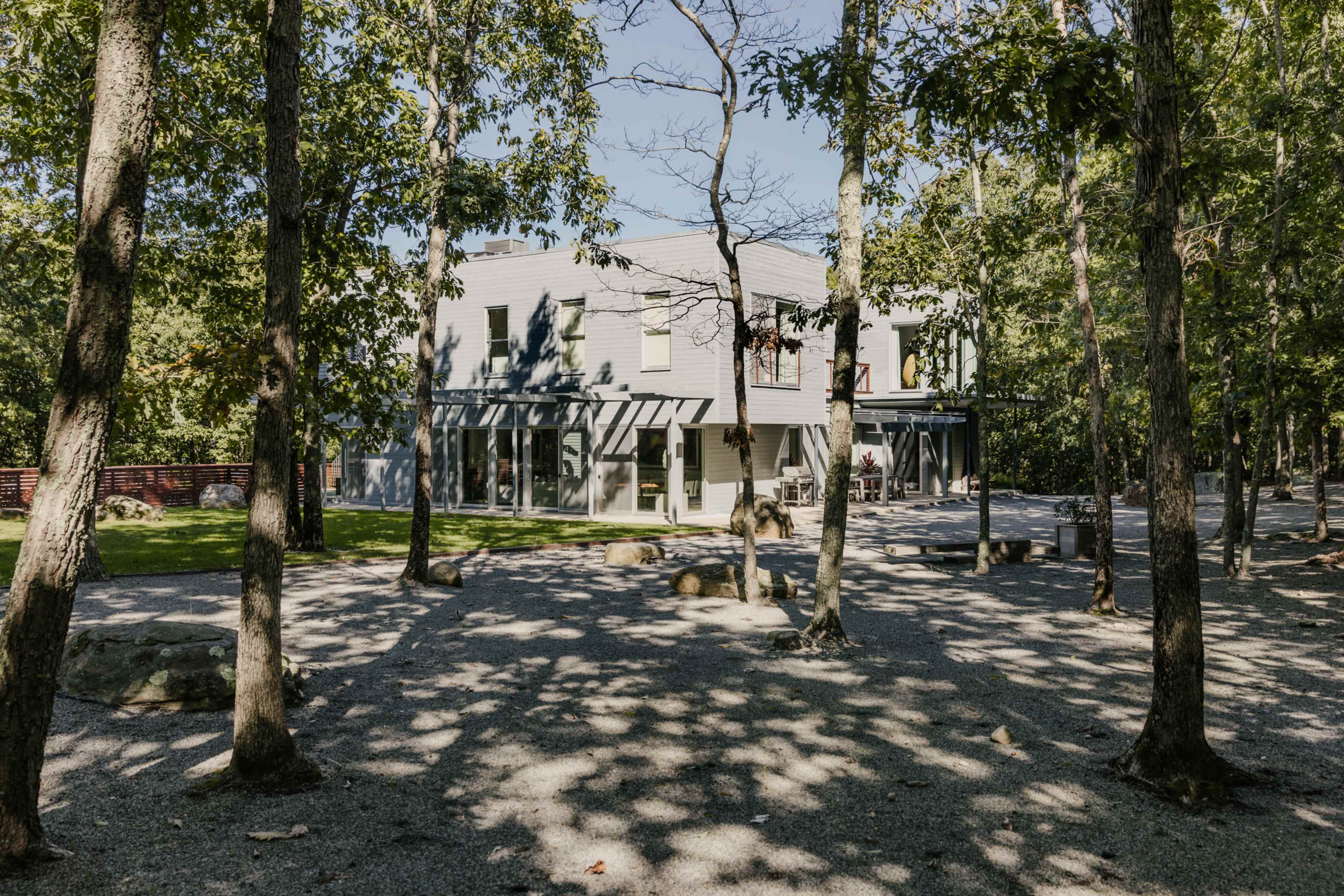 A modern two-story house is surrounded by trees and gravel pathways in a wooded area.