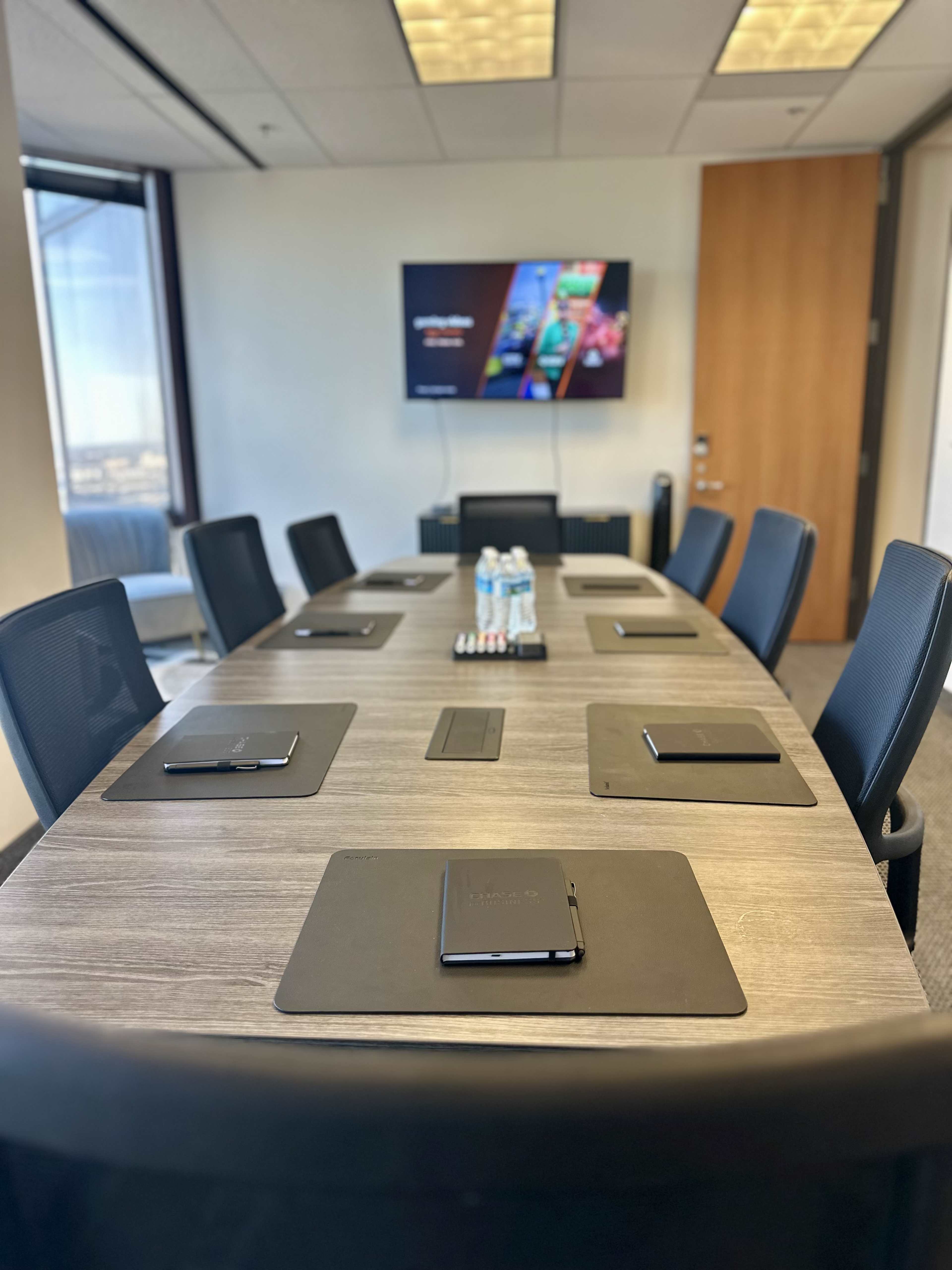 A modern conference room features a long wooden table with black notepads and pens at each seat, surrounded by ergonomic chairs, and is equipped with a wall-mounted screen displaying a presentation.