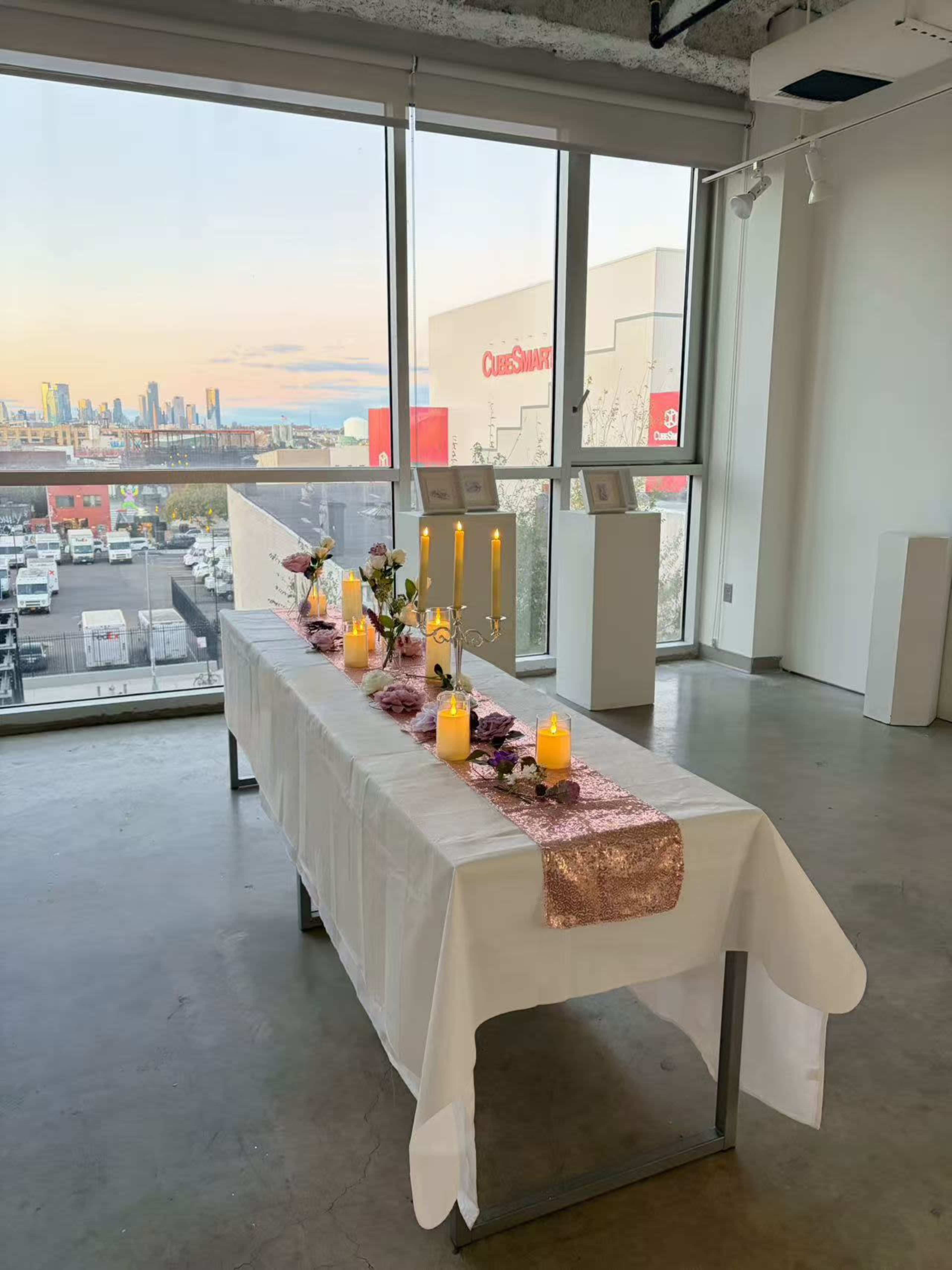 A rectangular table is set with candles, flowers, and a decorative runner, overlooking a city skyline through large windows.