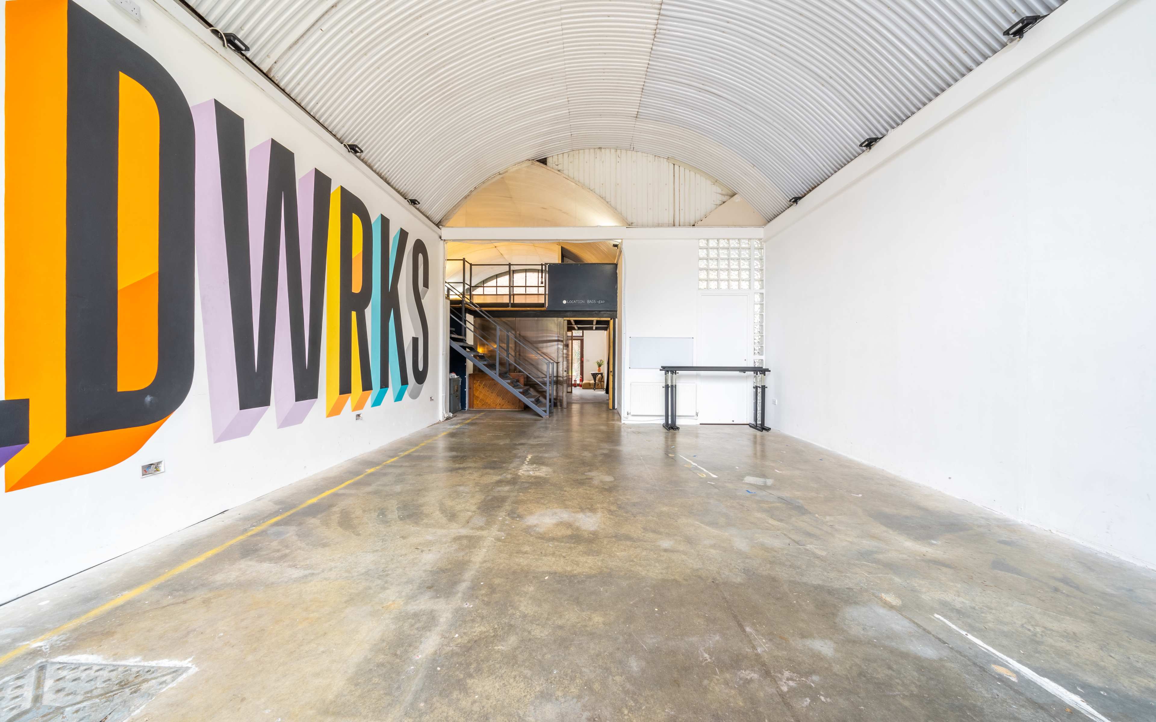 A spacious, industrial-style interior featuring a large mural that reads "LDWRKS" on one wall, with a staircase and an open area on the left.