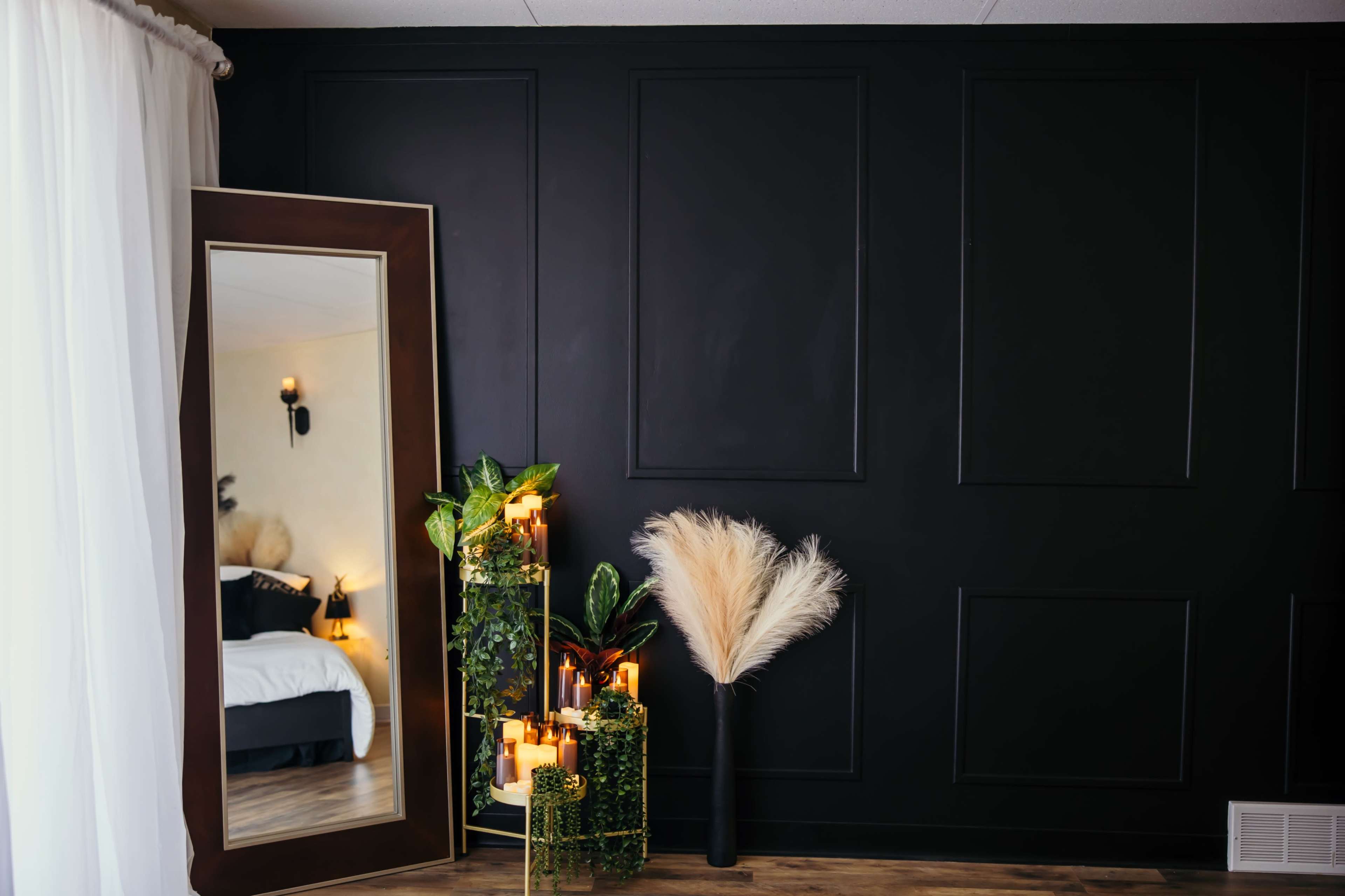The image features a stylish interior with a large mirror reflecting a bed and decorative plants alongside candles and a pampas grass arrangement against a dark wall.