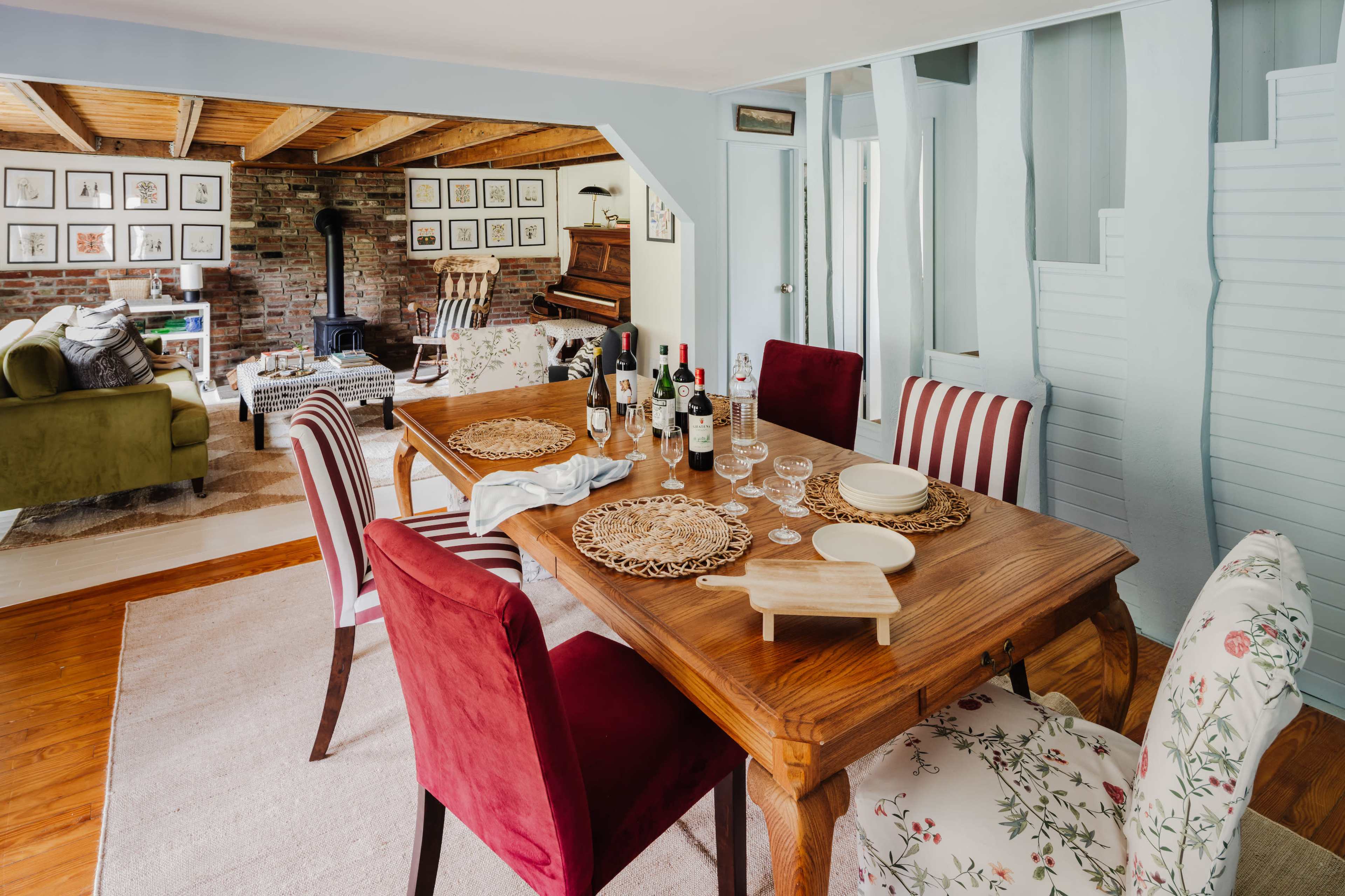 A wooden dining table is set with plates and glasses, surrounded by upholstered chairs, in a bright room that opens to a cozy living area with a piano and framed artwork.