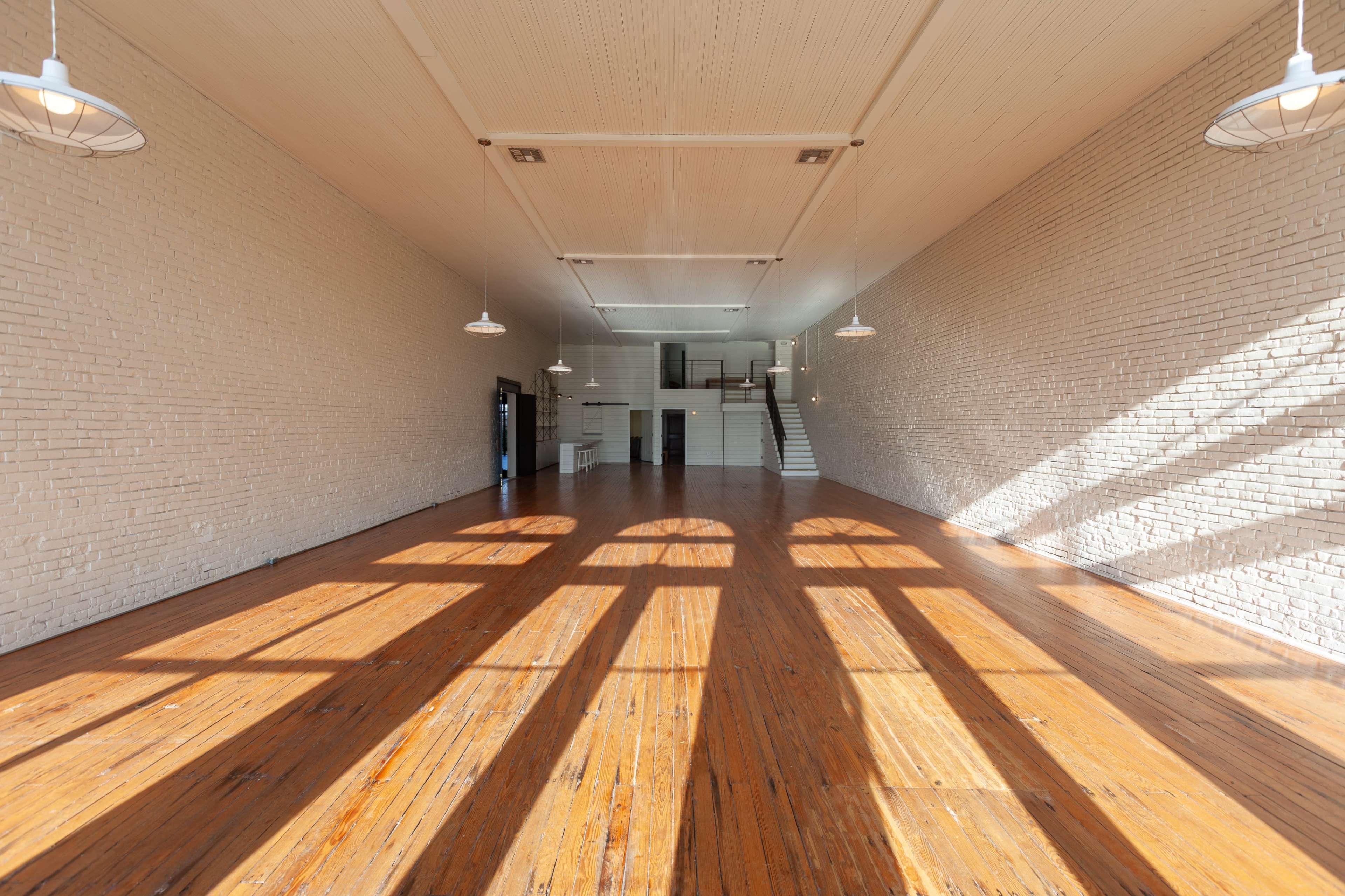 The image shows a spacious, unfurnished interior with wooden floors and large windows casting distinct shadows on the walls.