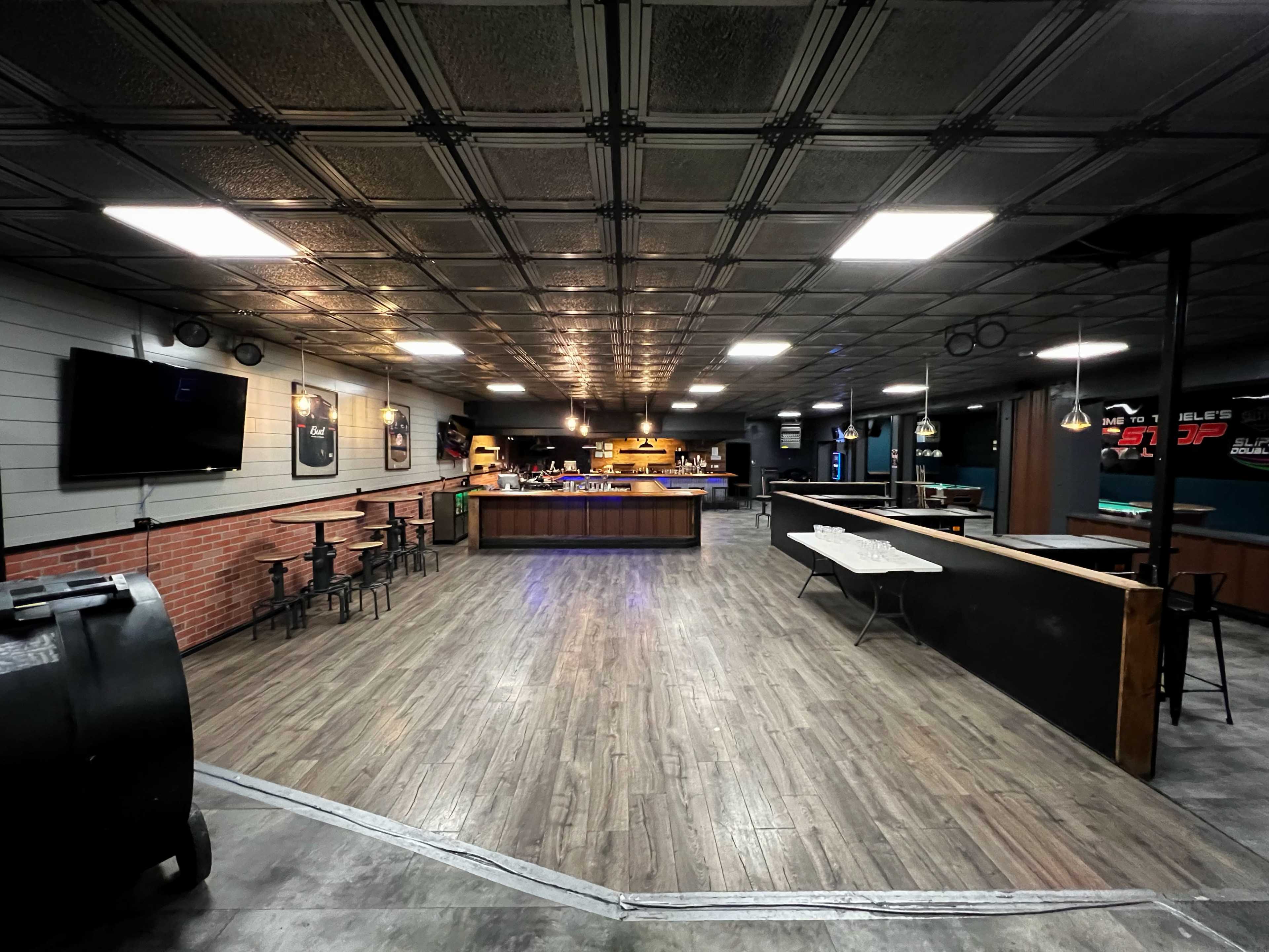 A spacious, dimly-lit interior of a bar or entertainment venue with a wooden bar counter, seating areas, and a patterned ceiling.