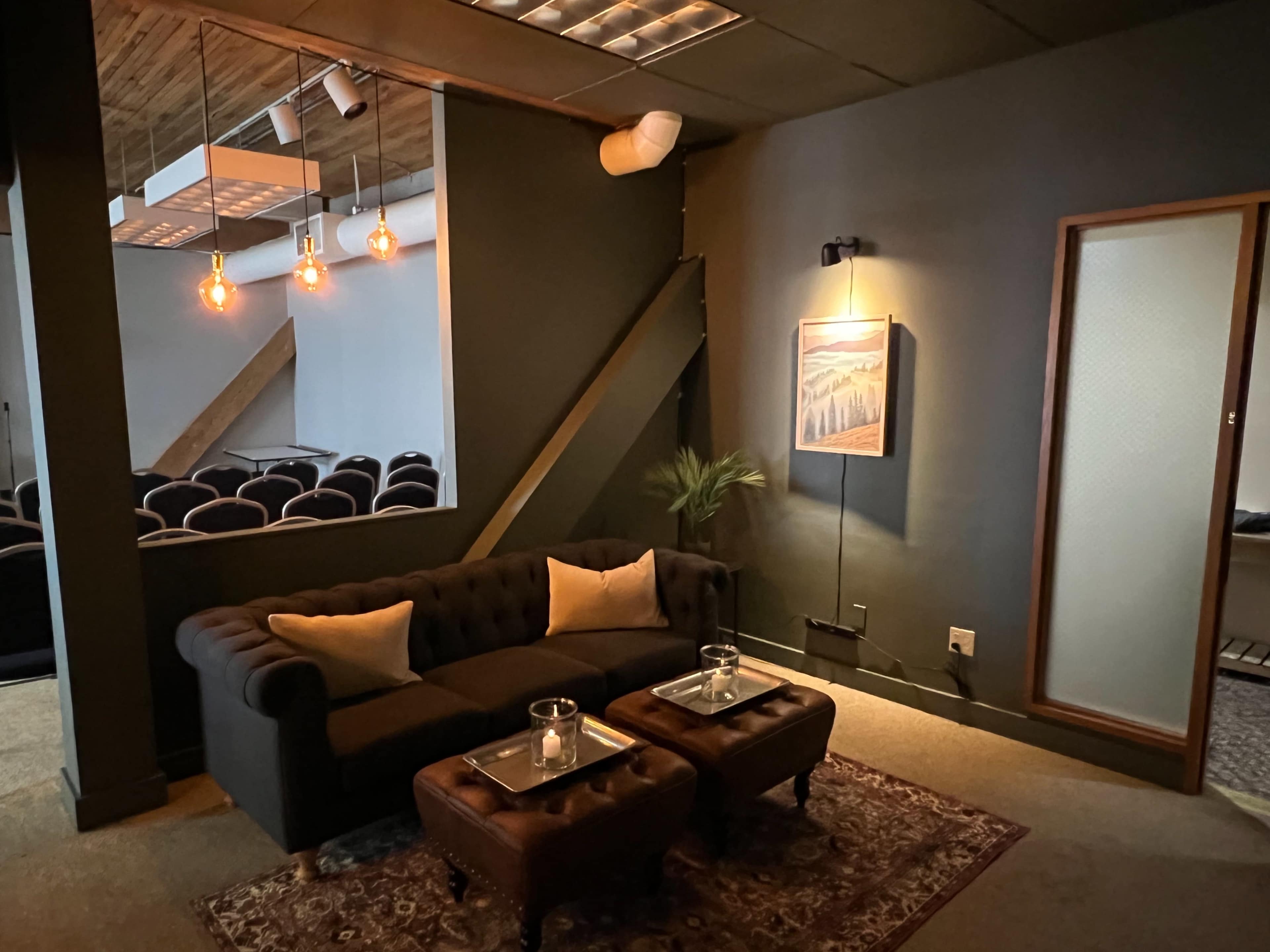 A cozy lounge area with a dark sofa, two small coffee tables, and warm lighting, adjacent to a seating area that features rows of chairs.