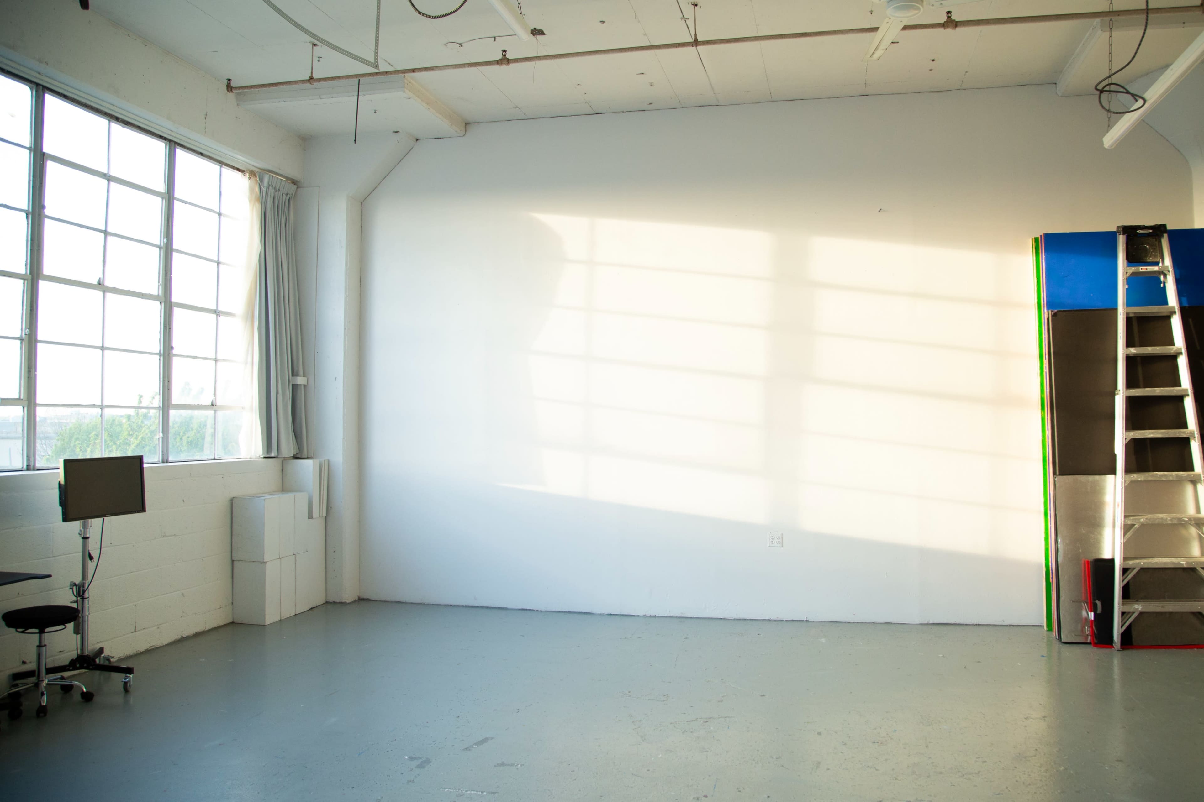 A well-lit studio space features a large window, a desk with a monitor, a ladder, and a blank white wall.