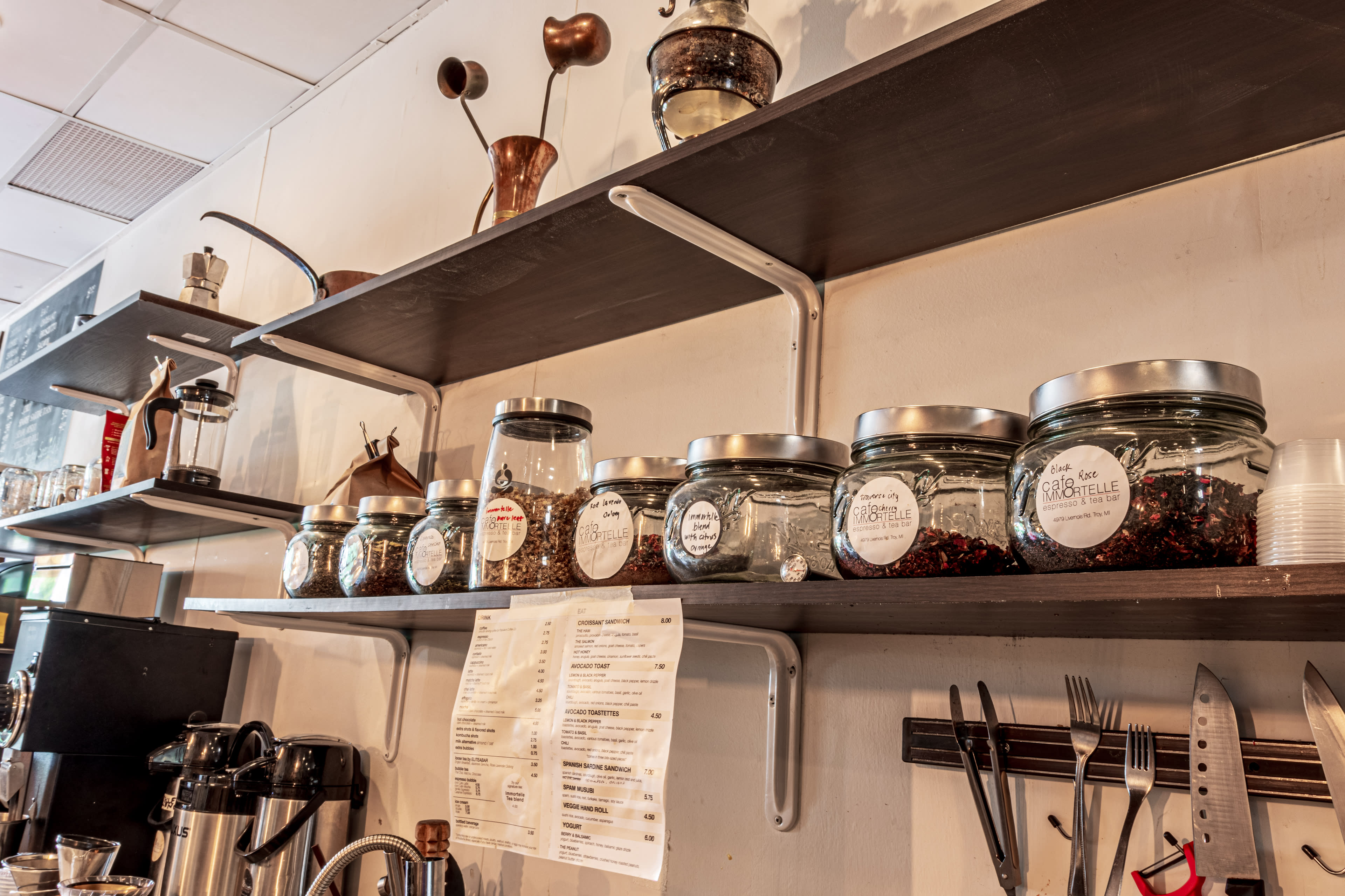 The image features a shelf in a café displaying glass jars filled with various ingredients alongside a menu and kitchen utensils.