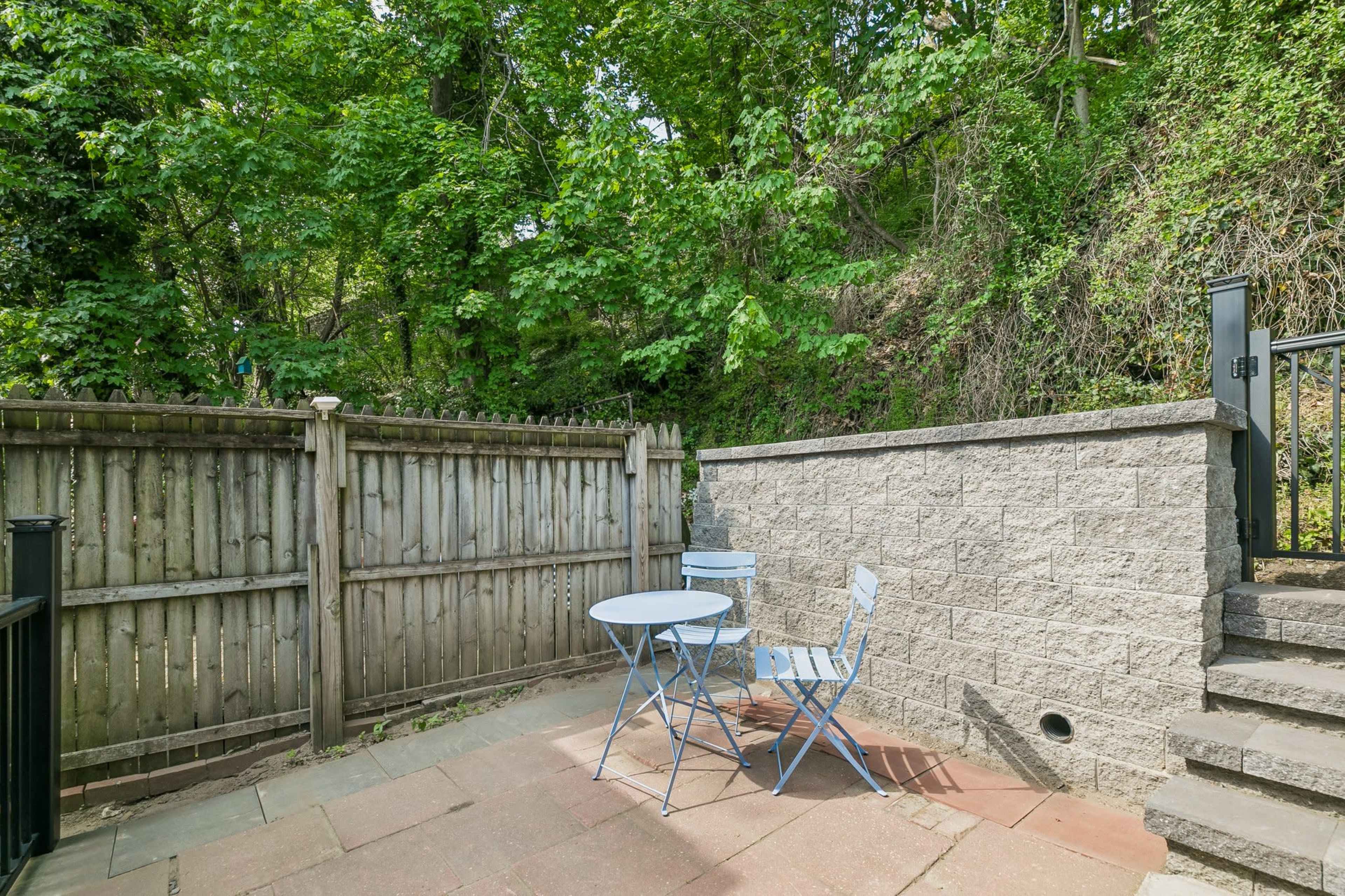 A small outdoor patio features a blue bistro table and two chairs, surrounded by greenery and a wooden fence.