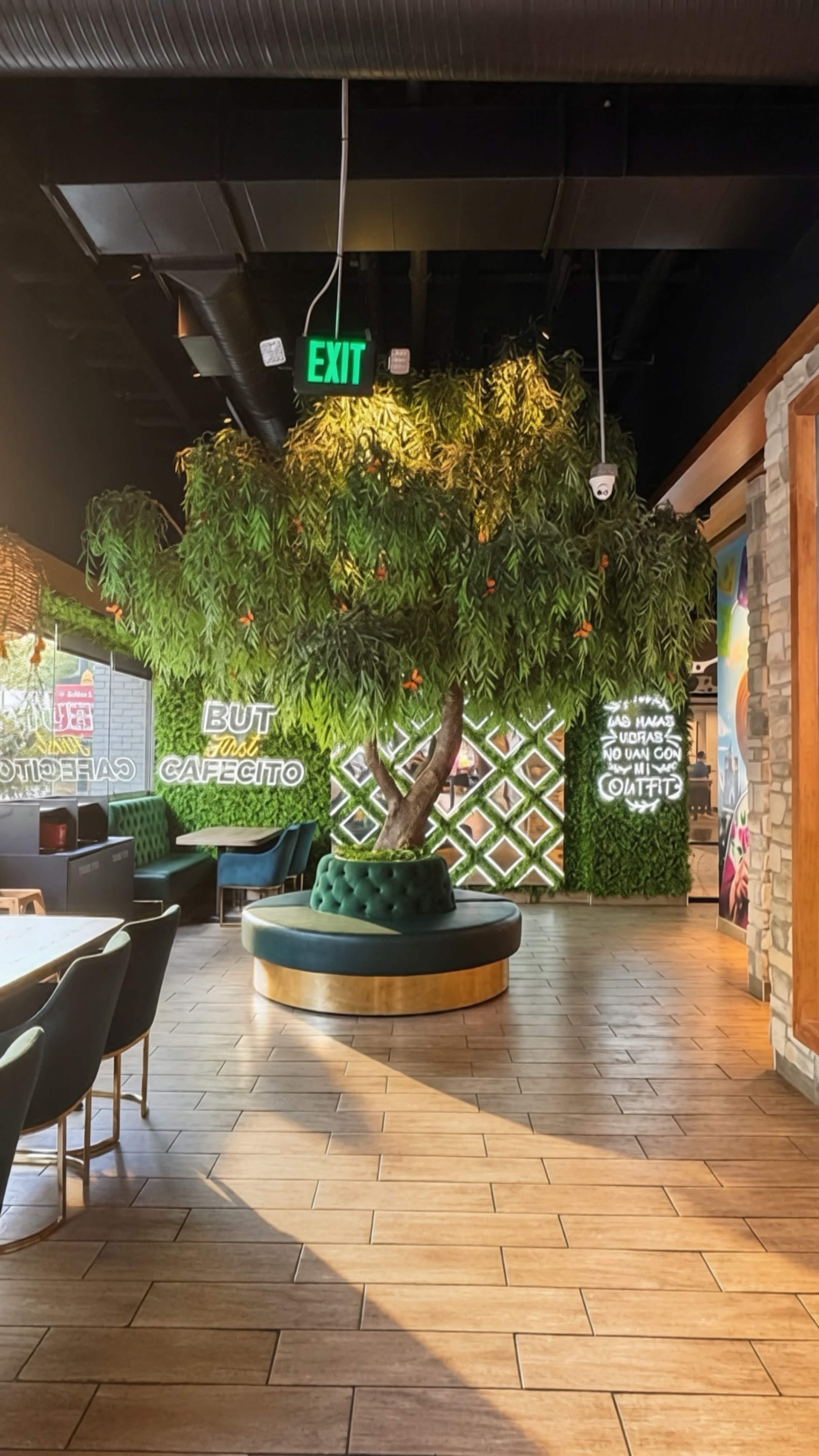 The interior of a restaurant features a large, lush tree with green leaves and orange accents, accompanied by a circular seating area and vibrant wall decor.