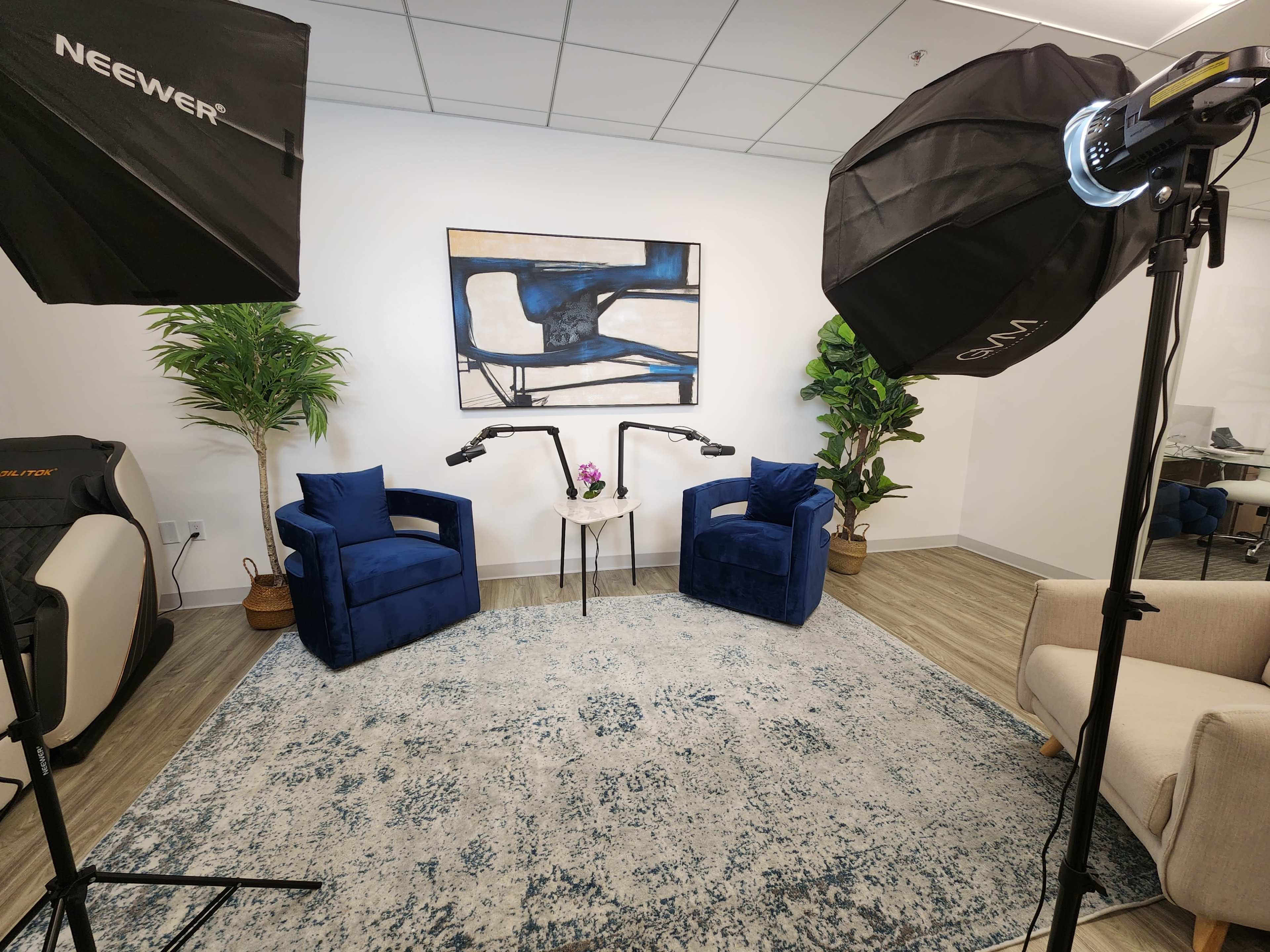 The image shows a small indoor setting with two blue chairs, a table with a plant, and photography lighting equipment.