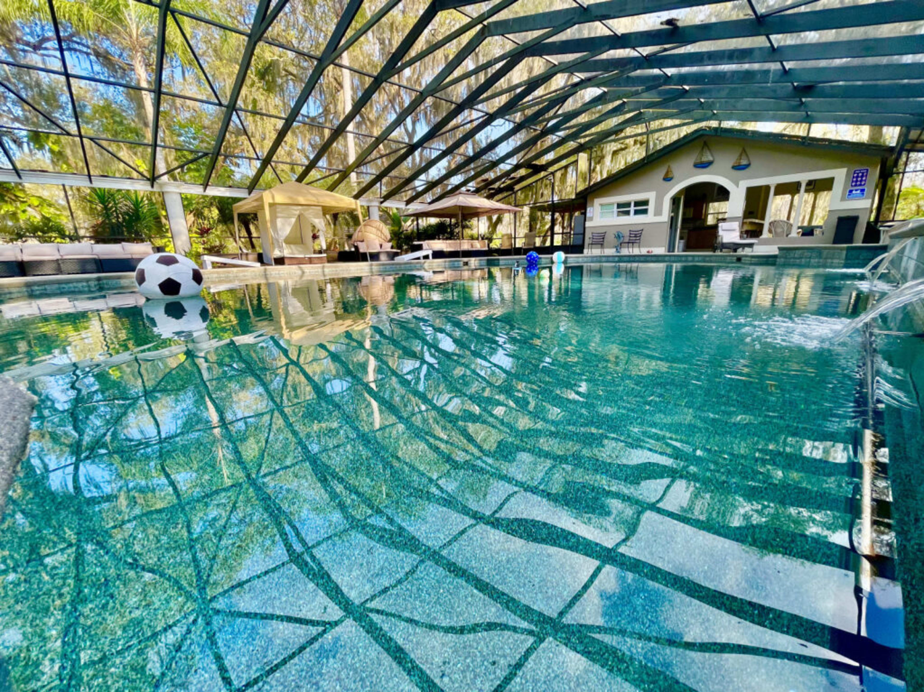 The image shows a spacious indoor pool area surrounded by a mesh enclosure, featuring clear water and various lounging amenities.