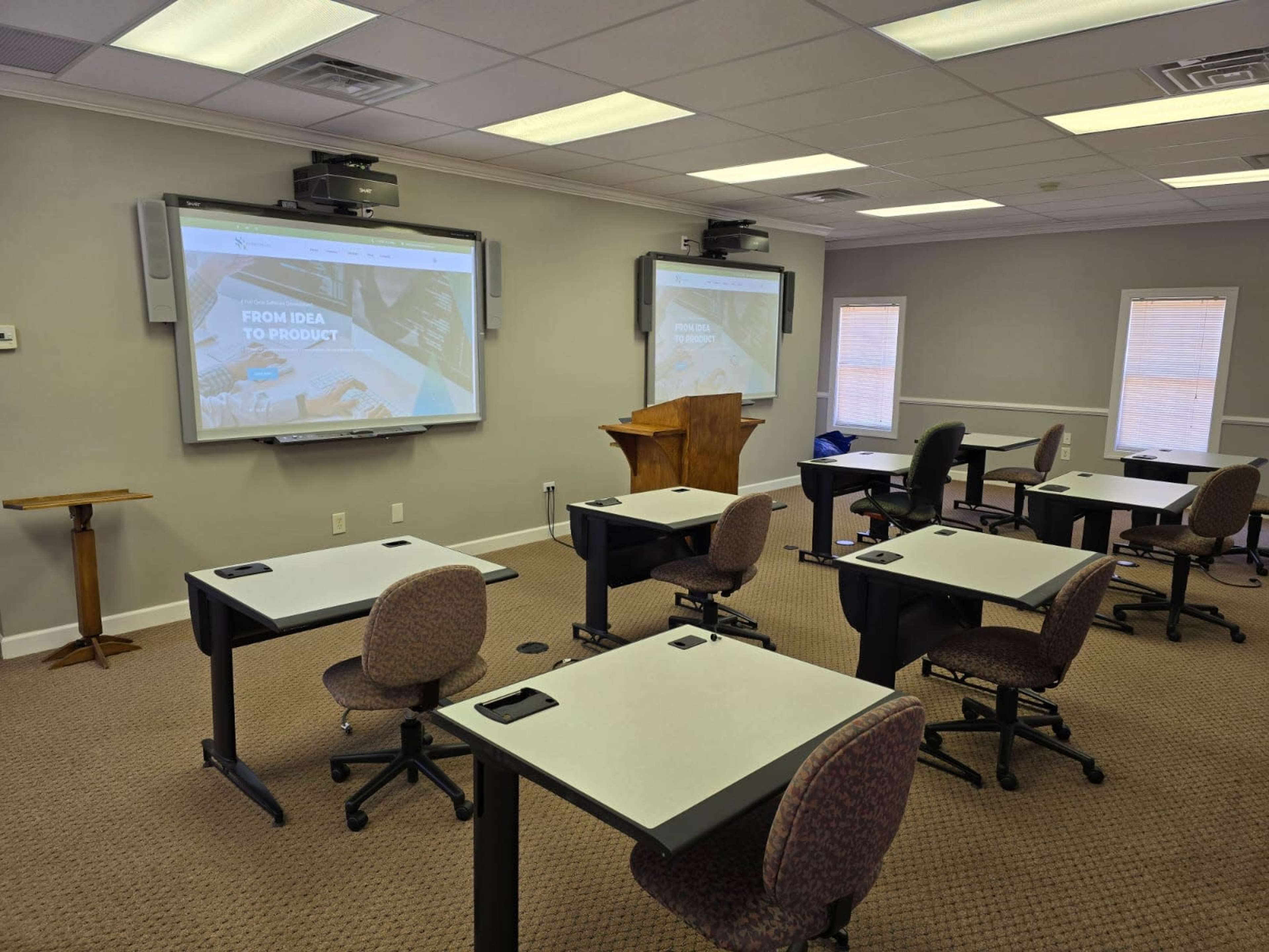 A classroom is set up with several desks facing two large screens displaying a presentation titled "FROM IDEA TO PRODUCT."