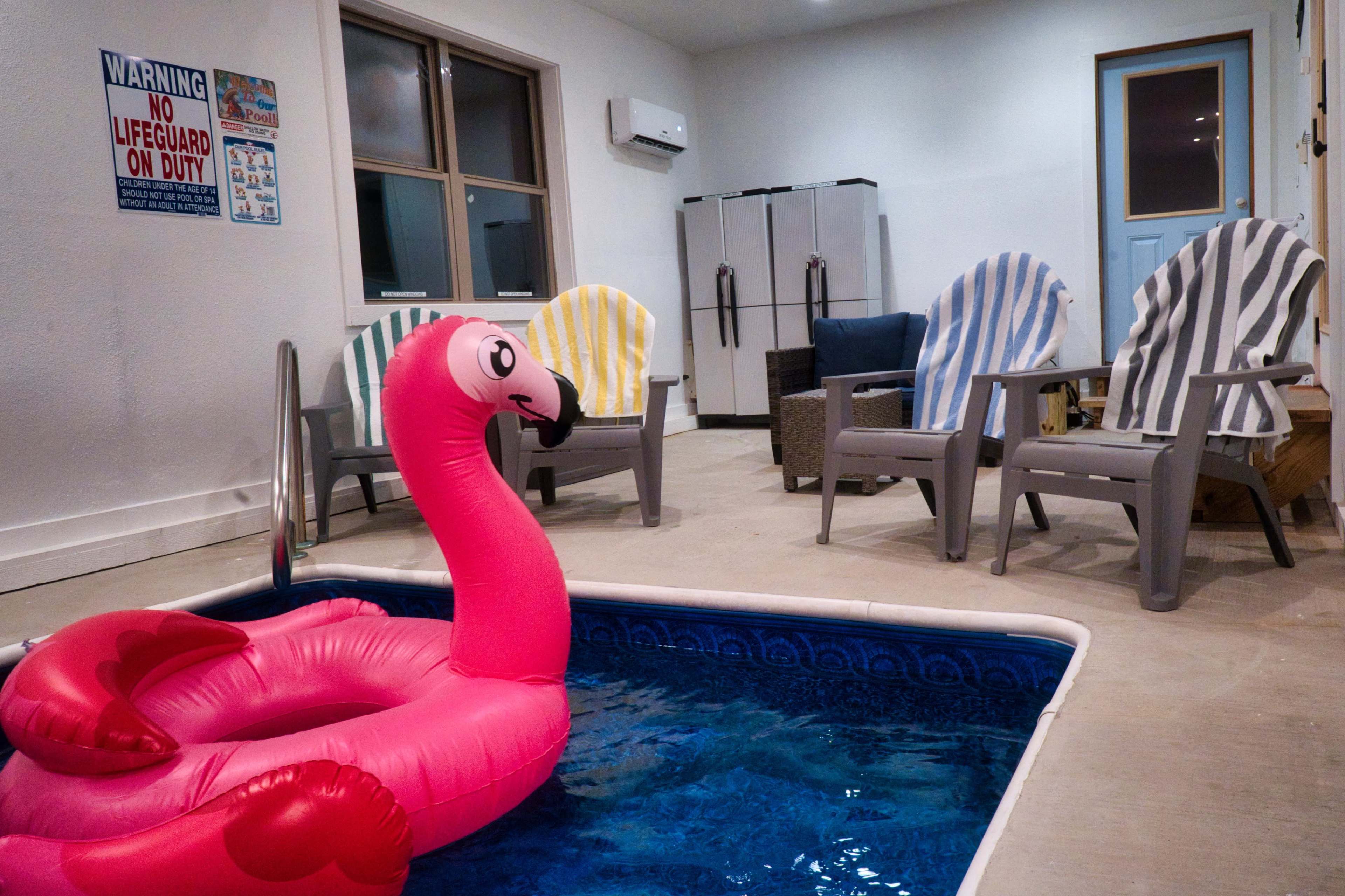 The image shows an indoor space featuring a small pool with a pink flamingo float, surrounded by colorful deck chairs and a set of refrigerators in the background.