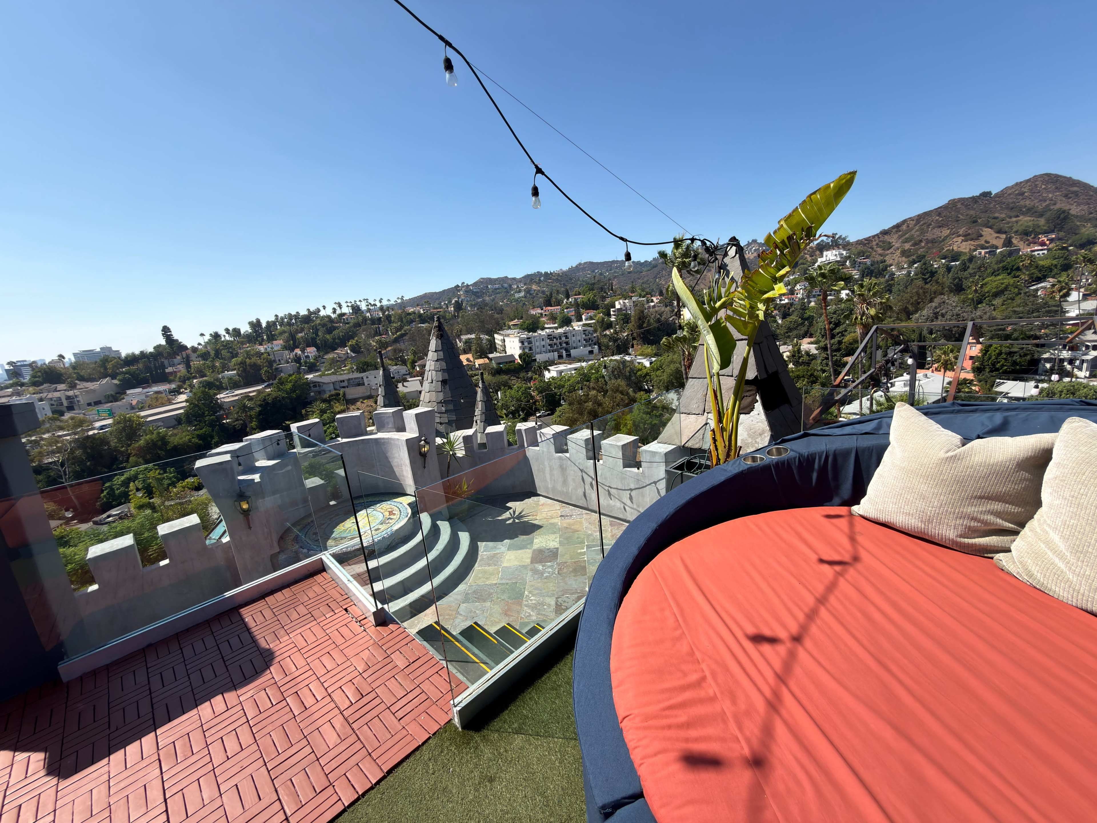 The image shows a rooftop terrace with a circular seating area, surrounded by a view of buildings and mountains in the background.