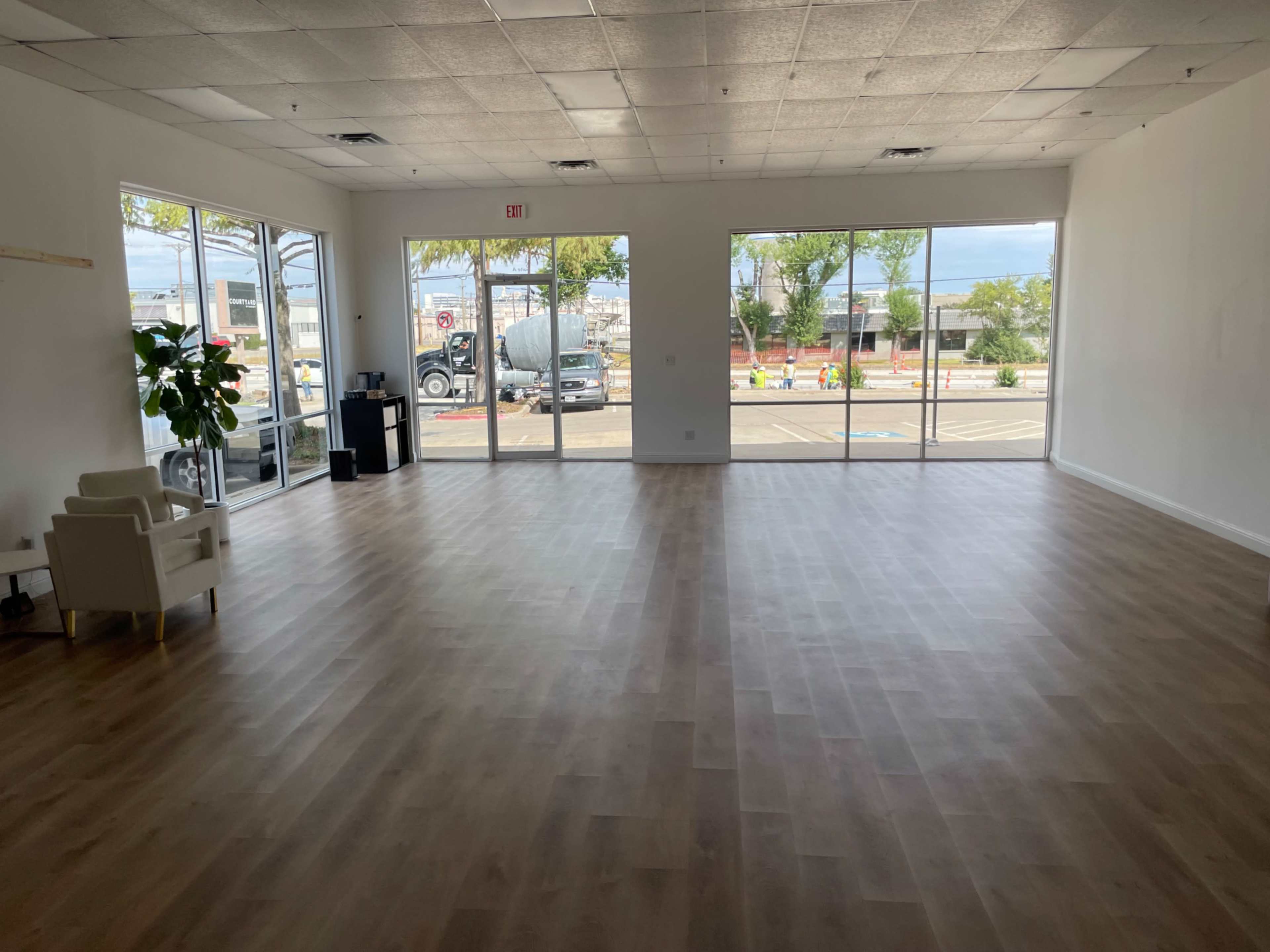 The image shows an empty commercial space with large windows, wooden flooring, and minimal furnishings.