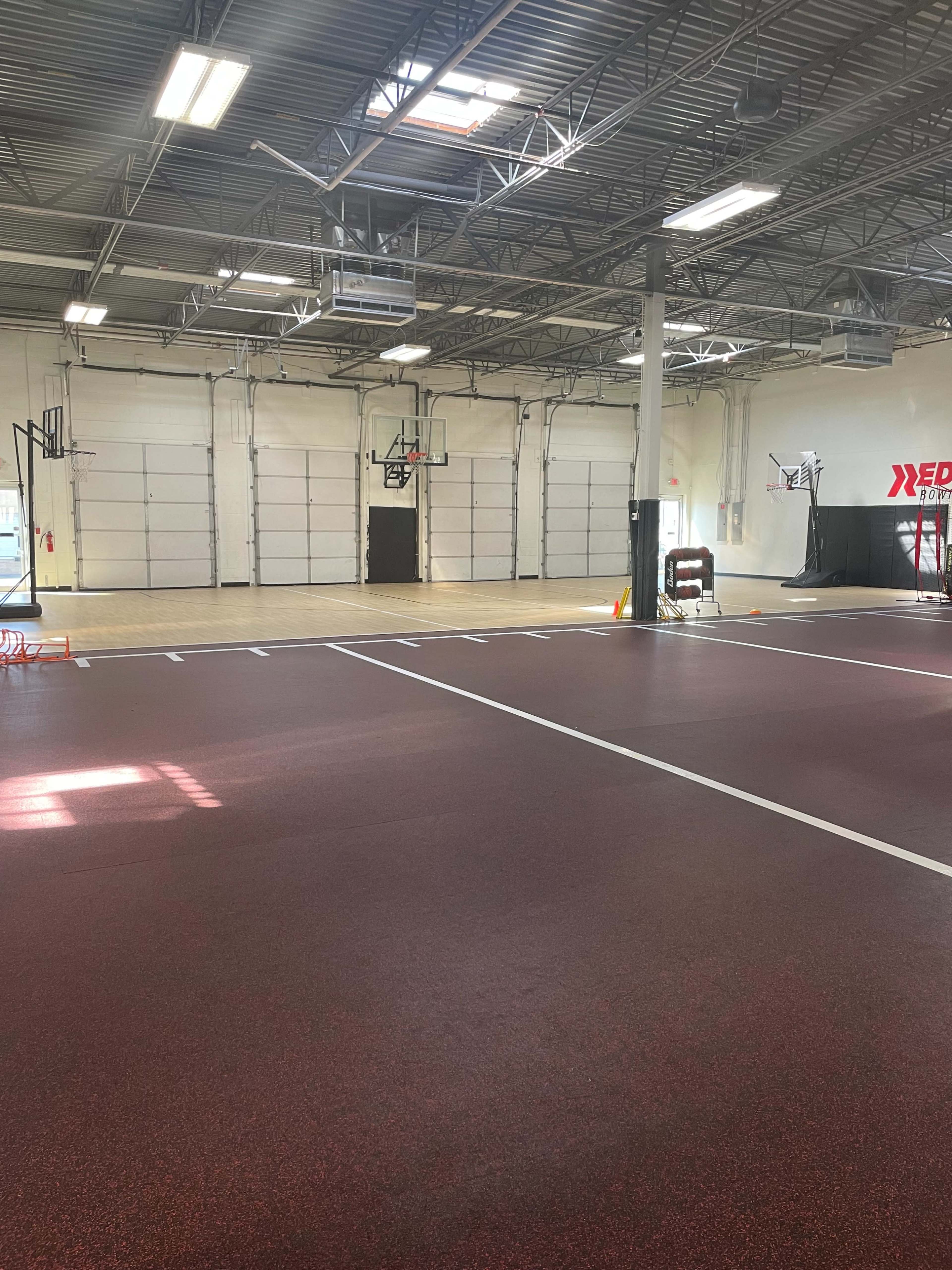 The image shows an indoor basketball court with a reddish floor, several basketball hoops, and a spacious, well-lit area.