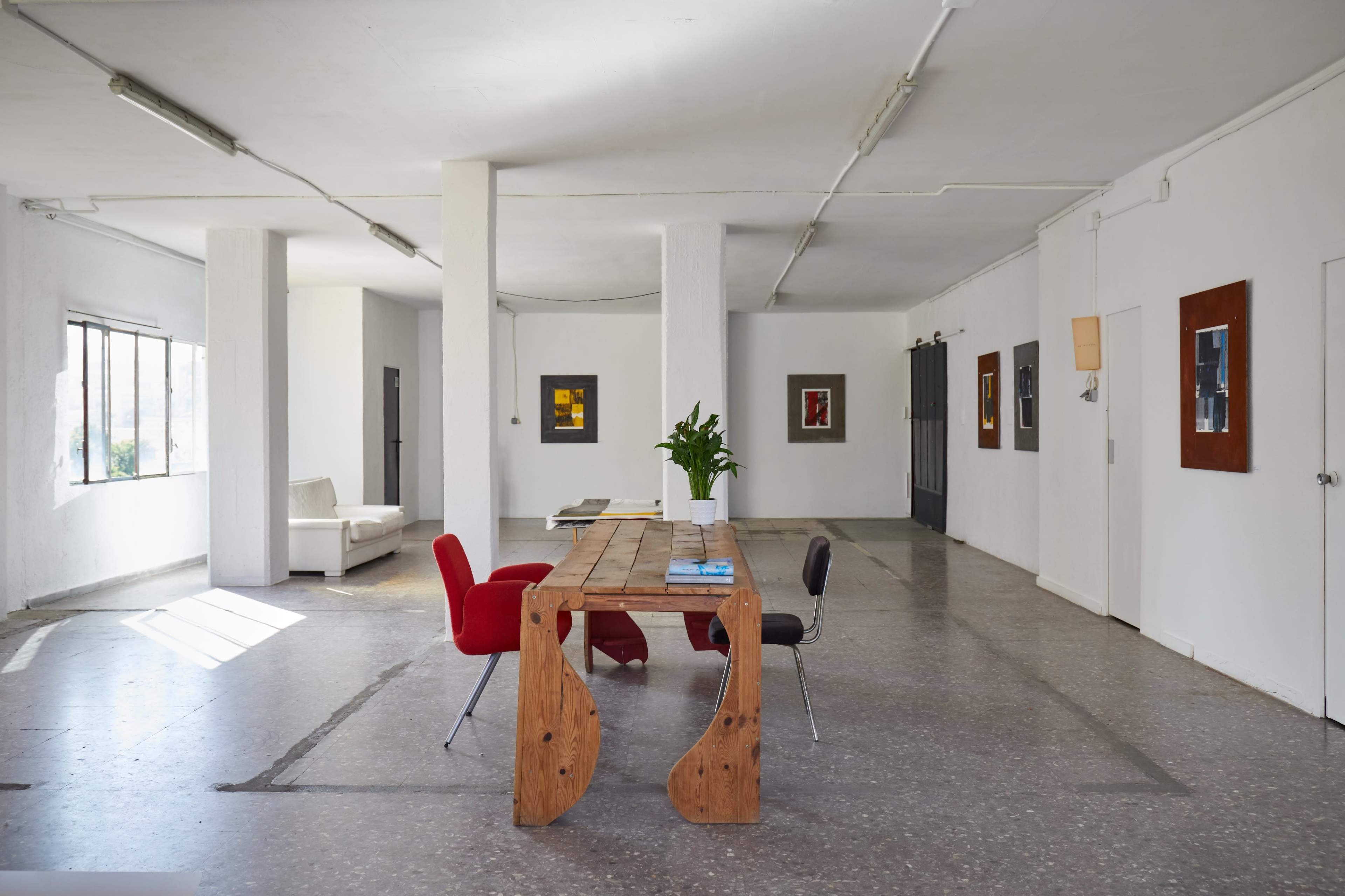 A spacious, minimalist room features a wooden table surrounded by chairs, with large windows allowing natural light and art pieces hanging on the walls.