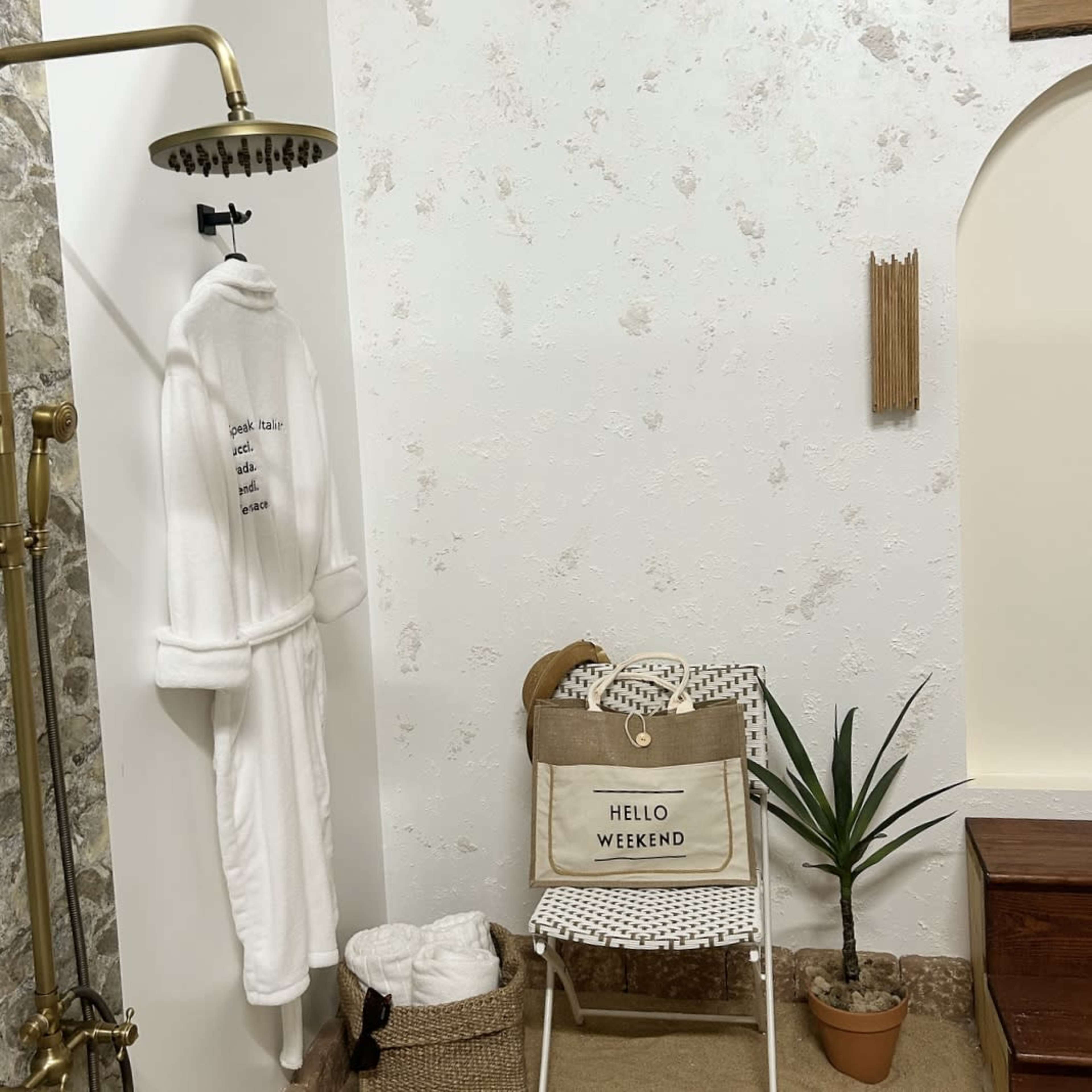 A bathroom corner features a plush white robe hanging next to a showerhead, a chair with a "HELLO WEEKEND" bag, a potted plant, and a basket of towels on the floor.