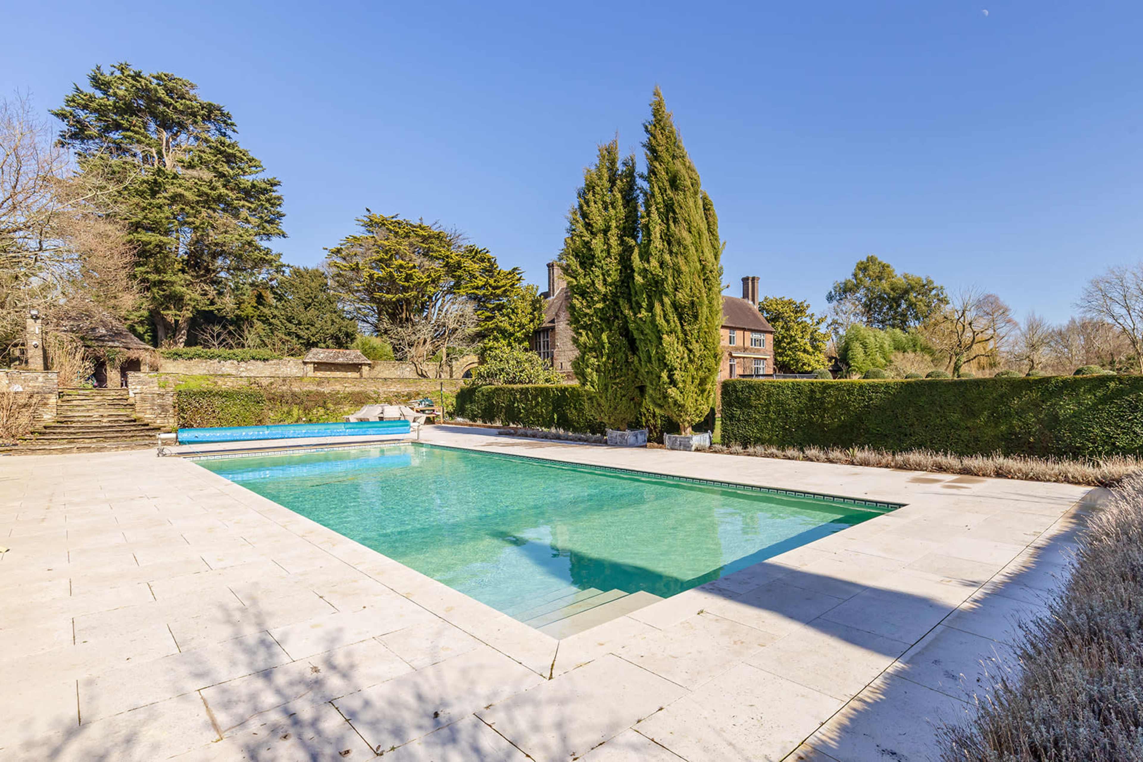 The image shows a rectangular swimming pool surrounded by stone paving and tall green trees, with a house visible in the background.
