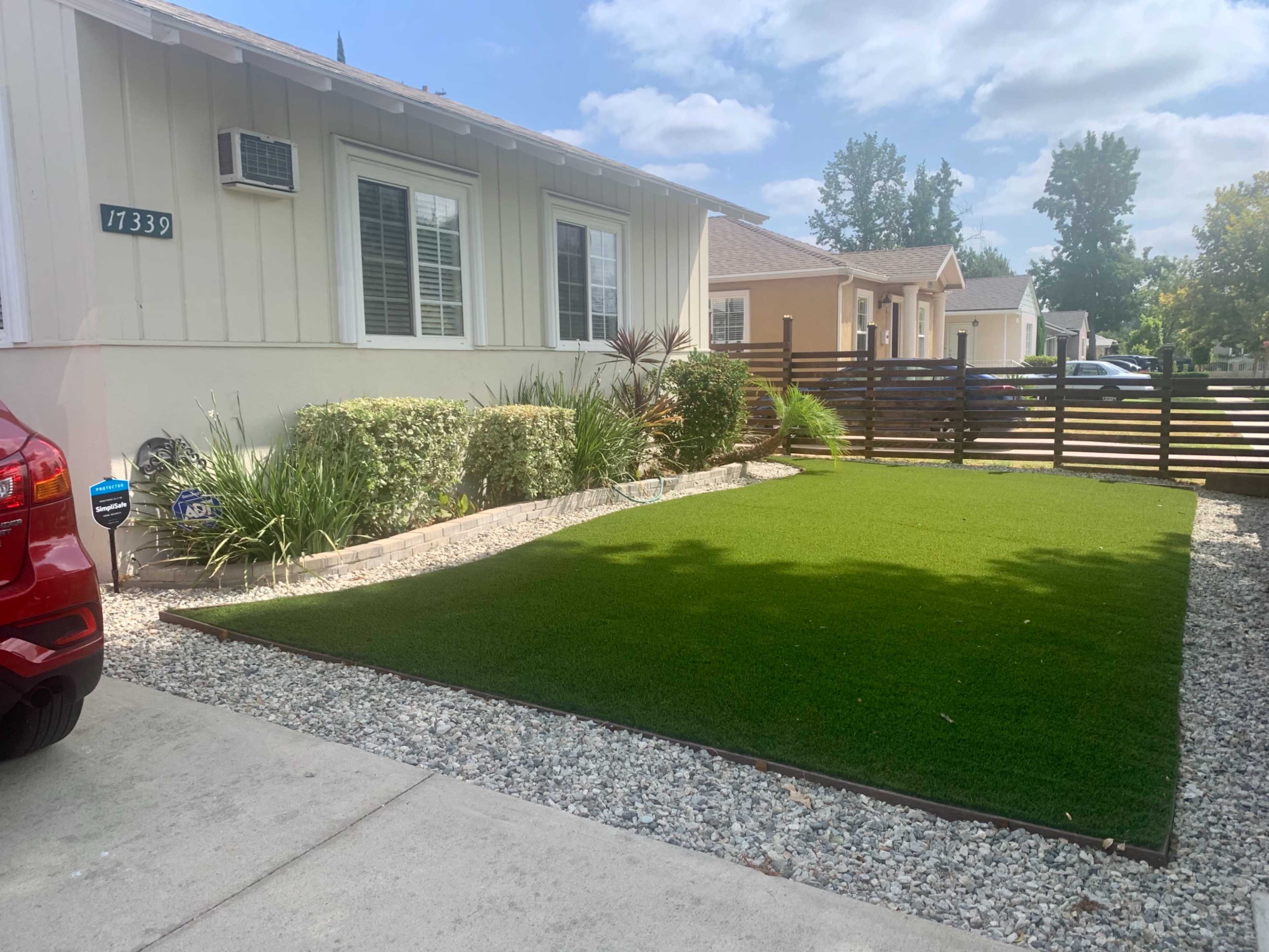 A residential front yard with artificial grass bordered by gravel and low shrubs, alongside a red car parked on the driveway.