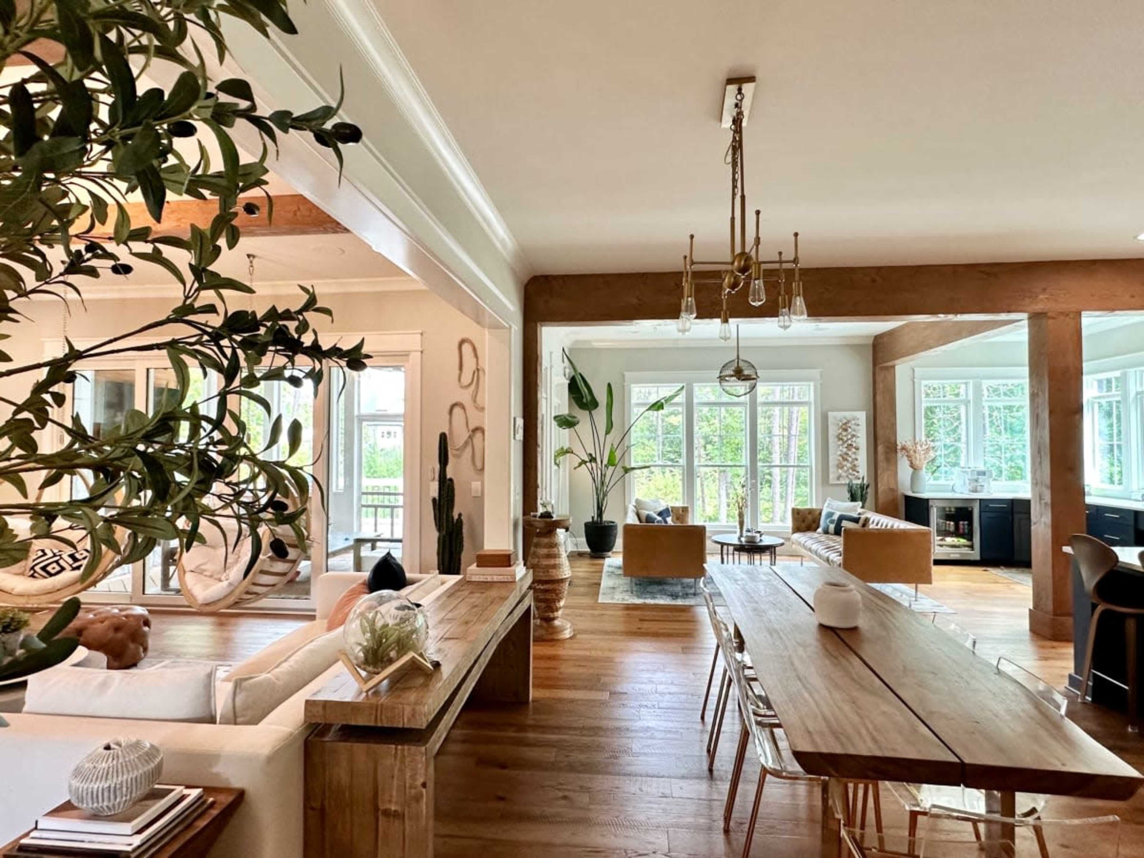A modern, open-plan living space featuring a wooden dining table, cozy seating areas, large windows, and greenery throughout.