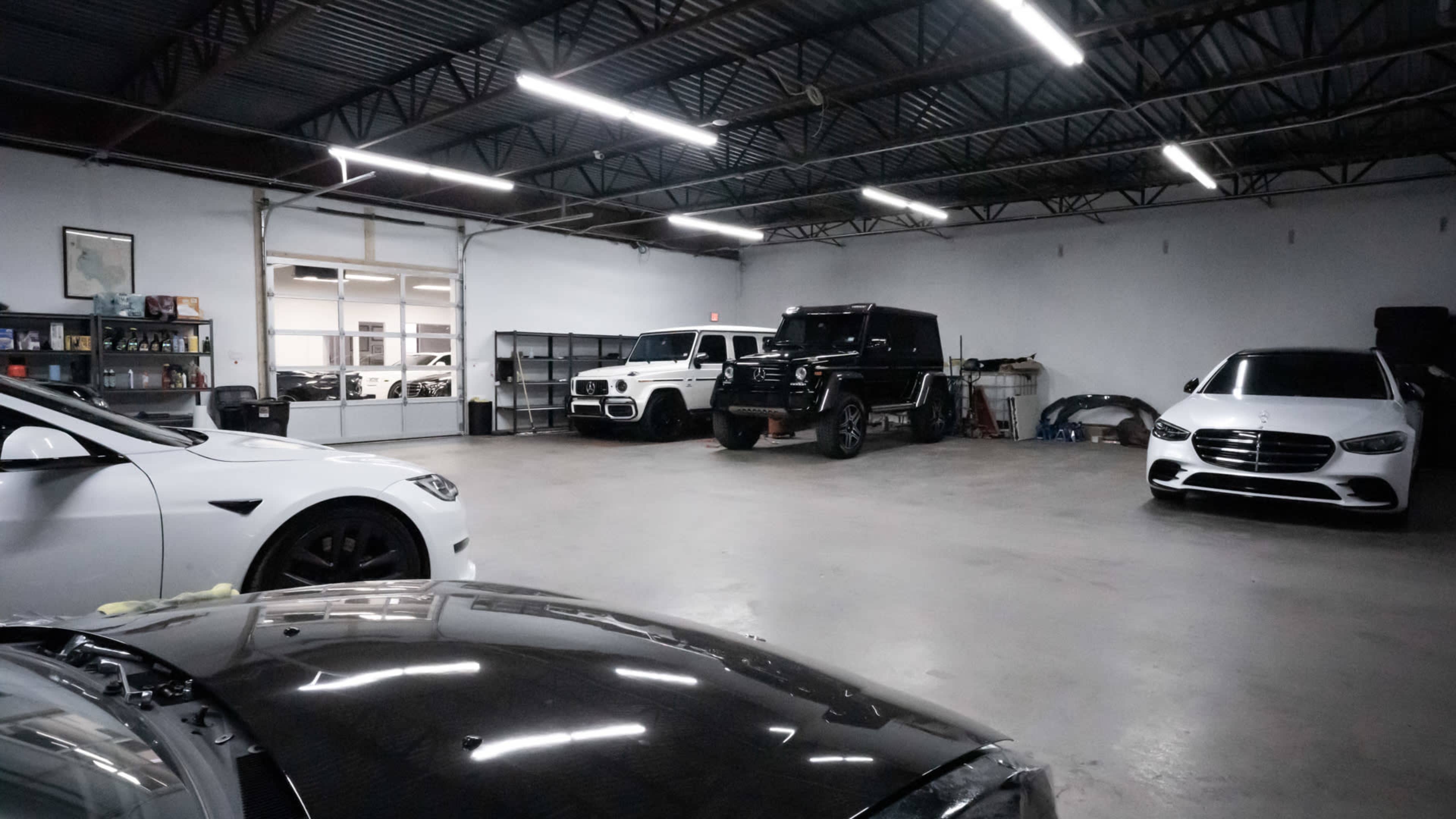 A spacious garage with several parked luxury cars, including a Tesla, a Mercedes, and a black SUV.