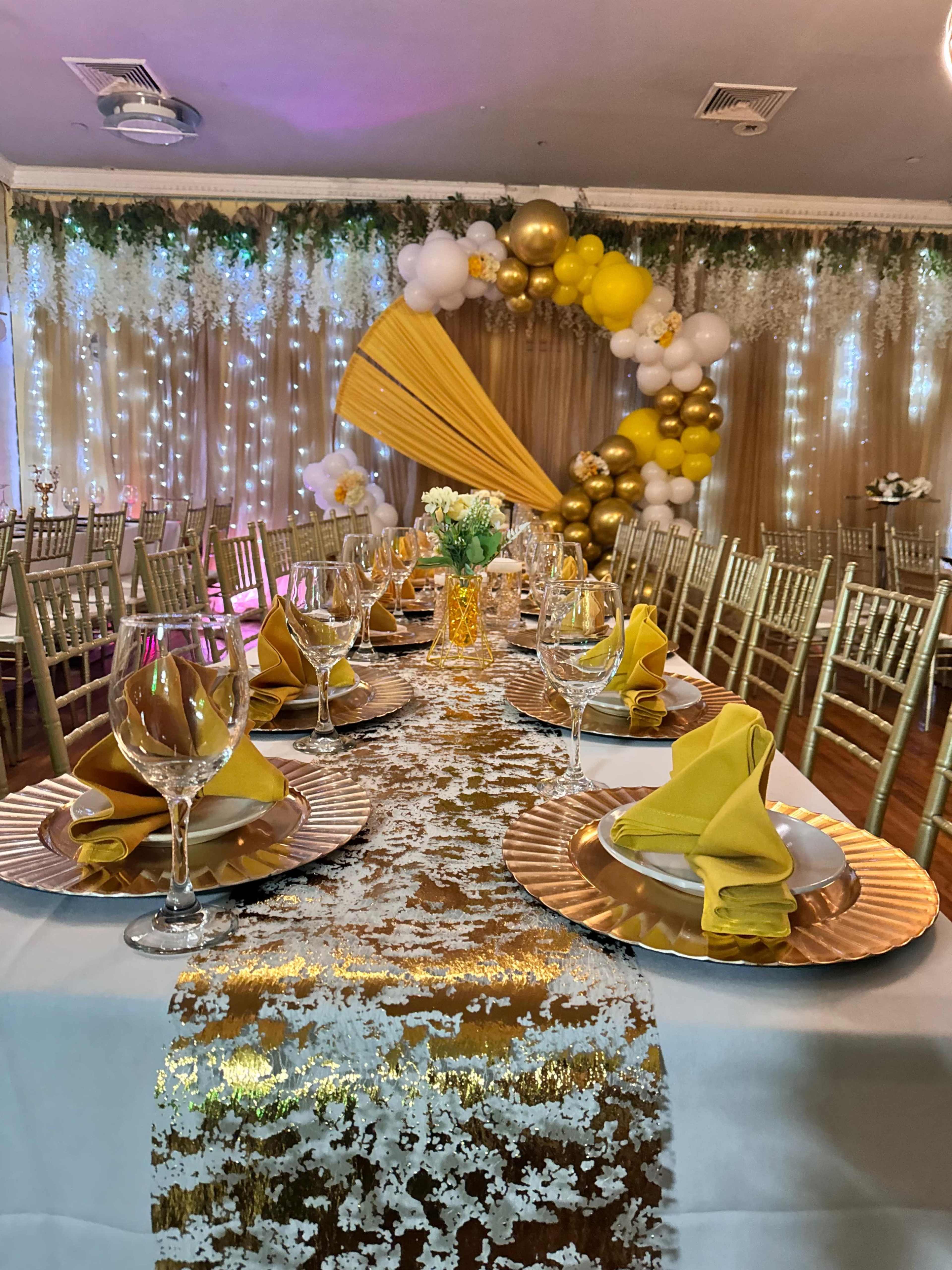 The image shows a decorated banquet table set with gold and yellow tableware, surrounded by elegant chairs, with a backdrop of draped fabric and balloons.