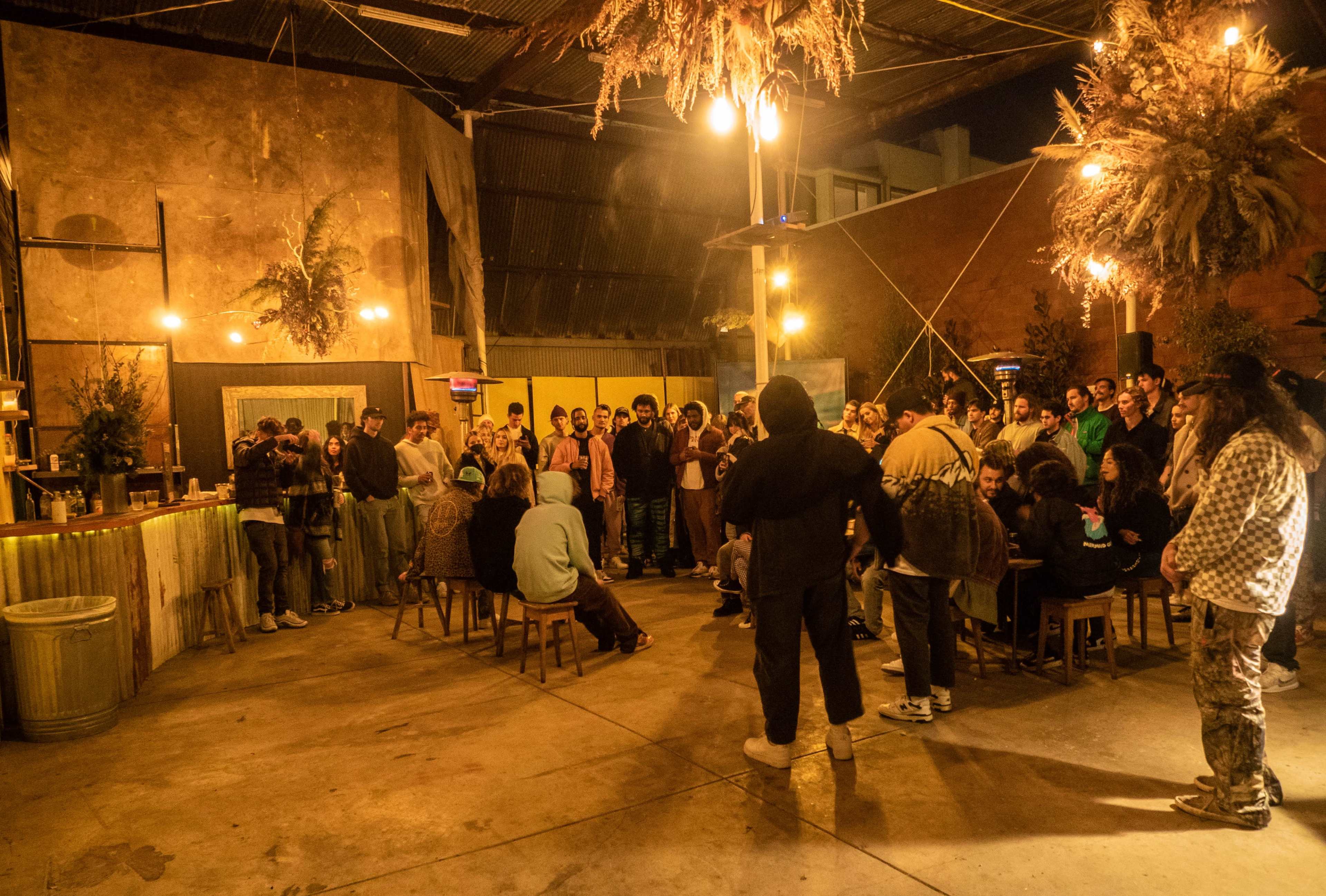 A crowd of people gathers in a dimly lit indoor space with rustic decor, wooden furniture, and hanging plants.
