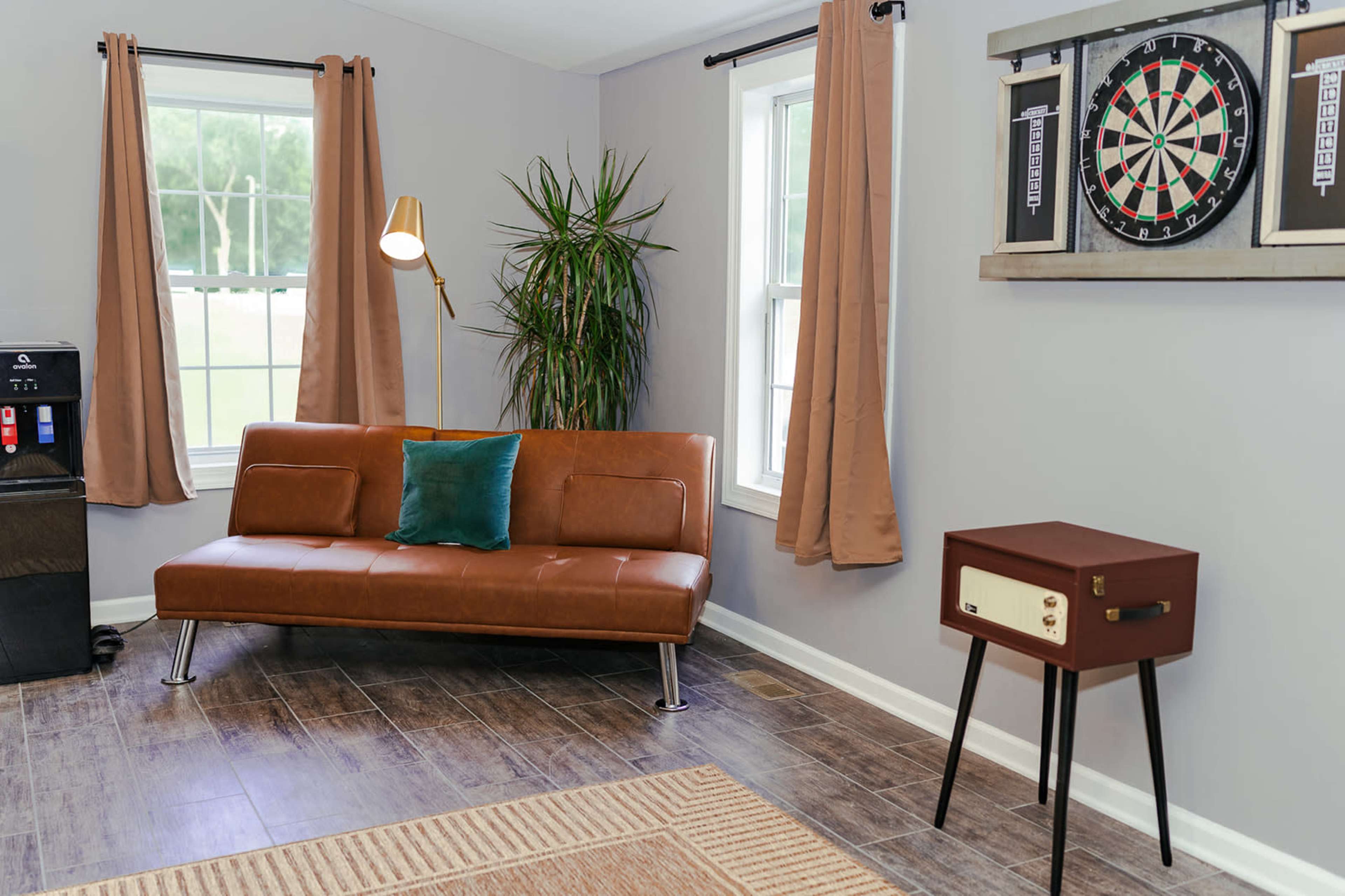 A living room with a brown sofa, a potted plant, a dartboard on the wall, and a vintage-style radio cabinet beside it.