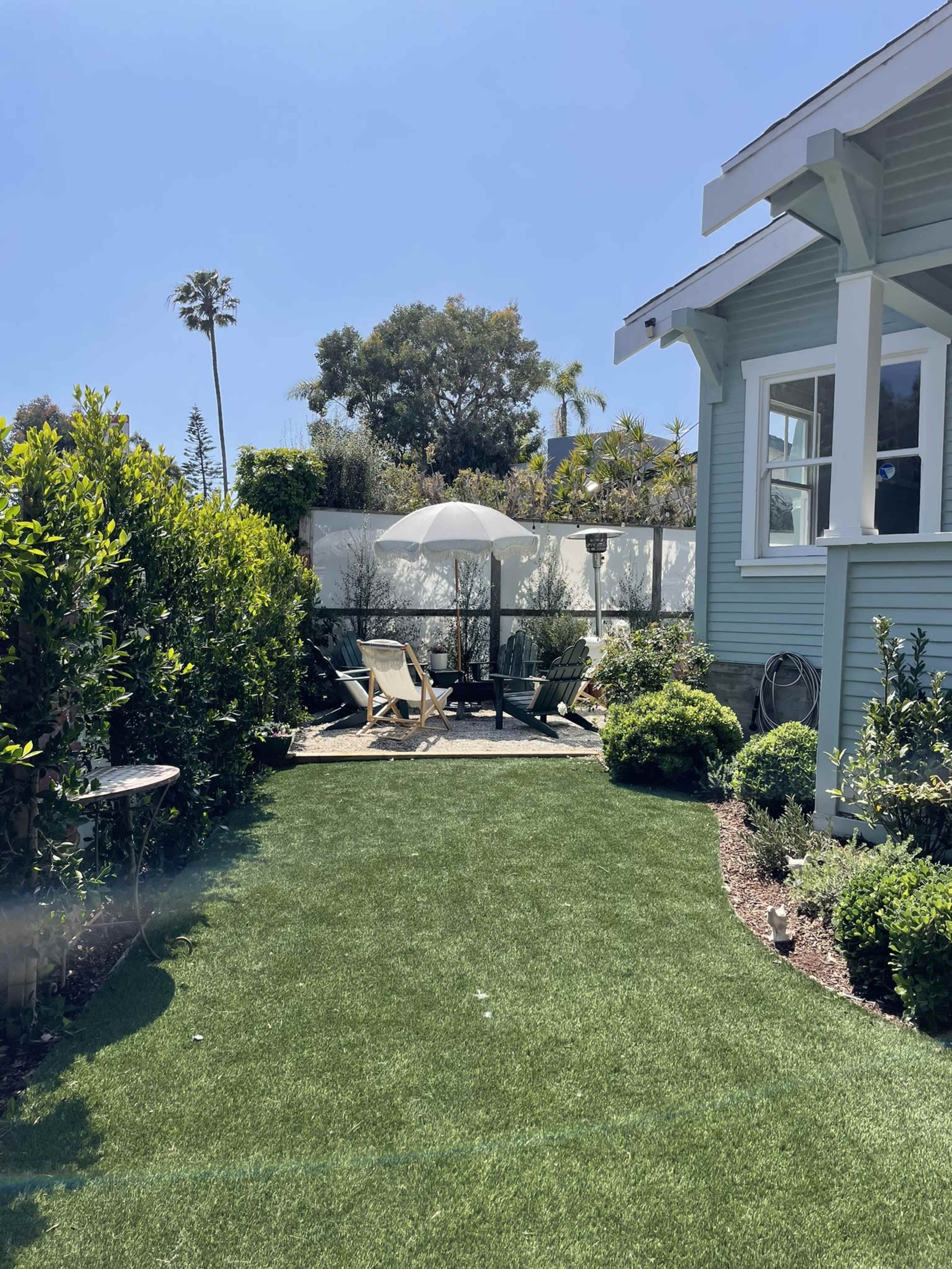 A sunny backyard garden with artificial grass, featuring a lounge chair, an umbrella, and neatly trimmed hedges.