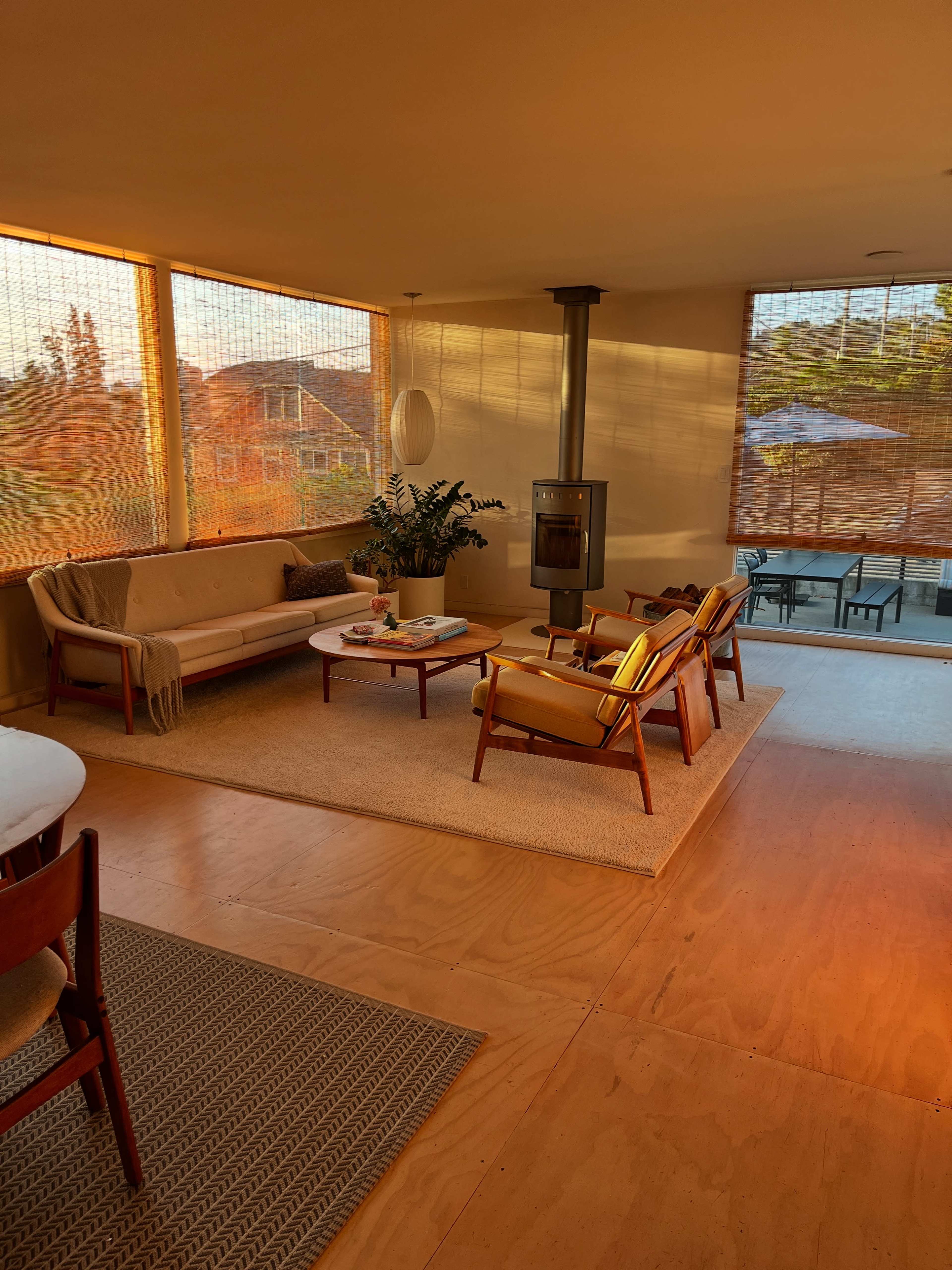 The image shows a light-filled living room featuring a seating area with two chairs and a sofa, a circular coffee table, and a wood stove, with large windows overlooking an outdoor patio.