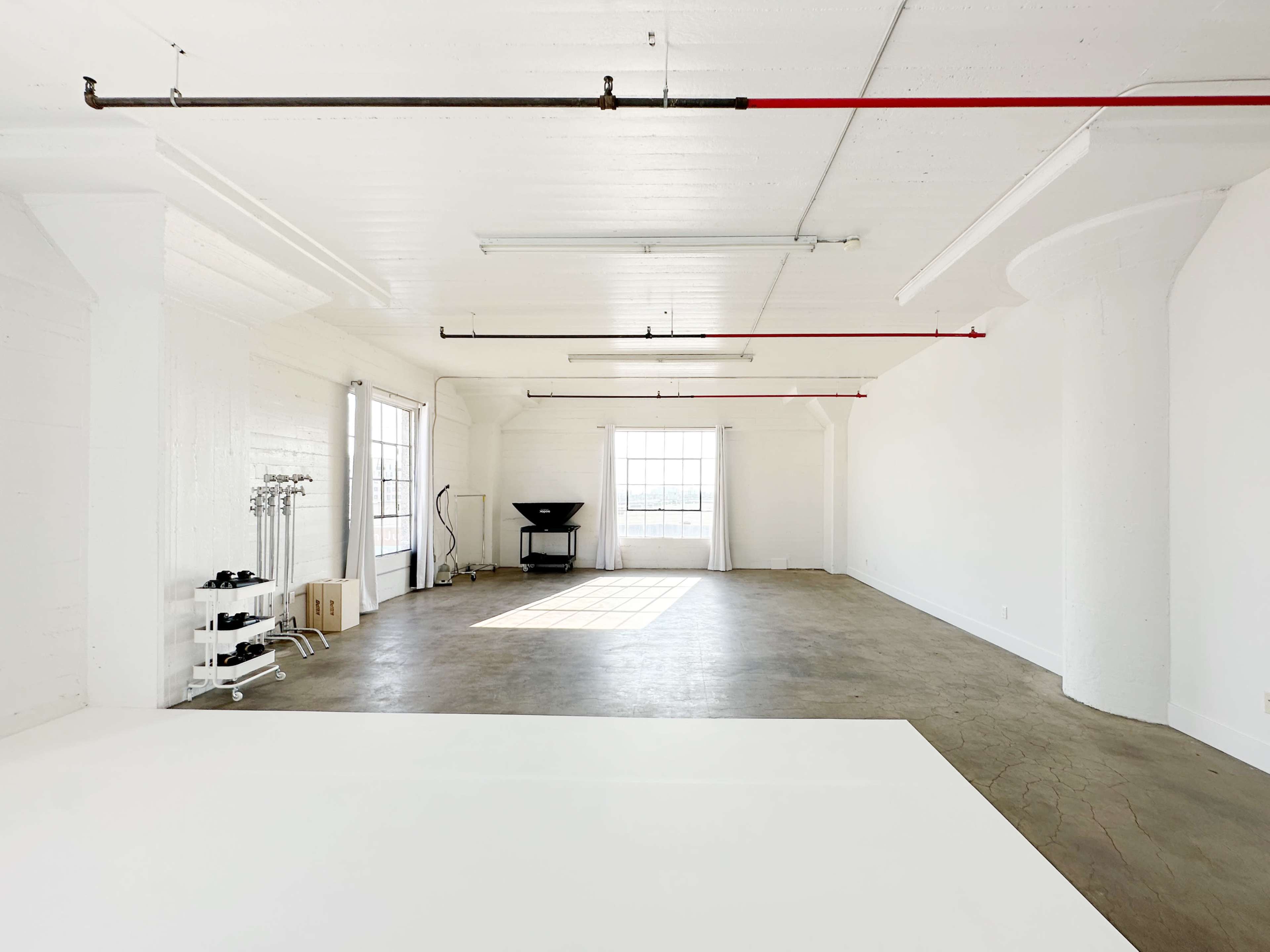 A spacious, empty room with white walls, large windows, and industrial piping, featuring minimal furnishings.