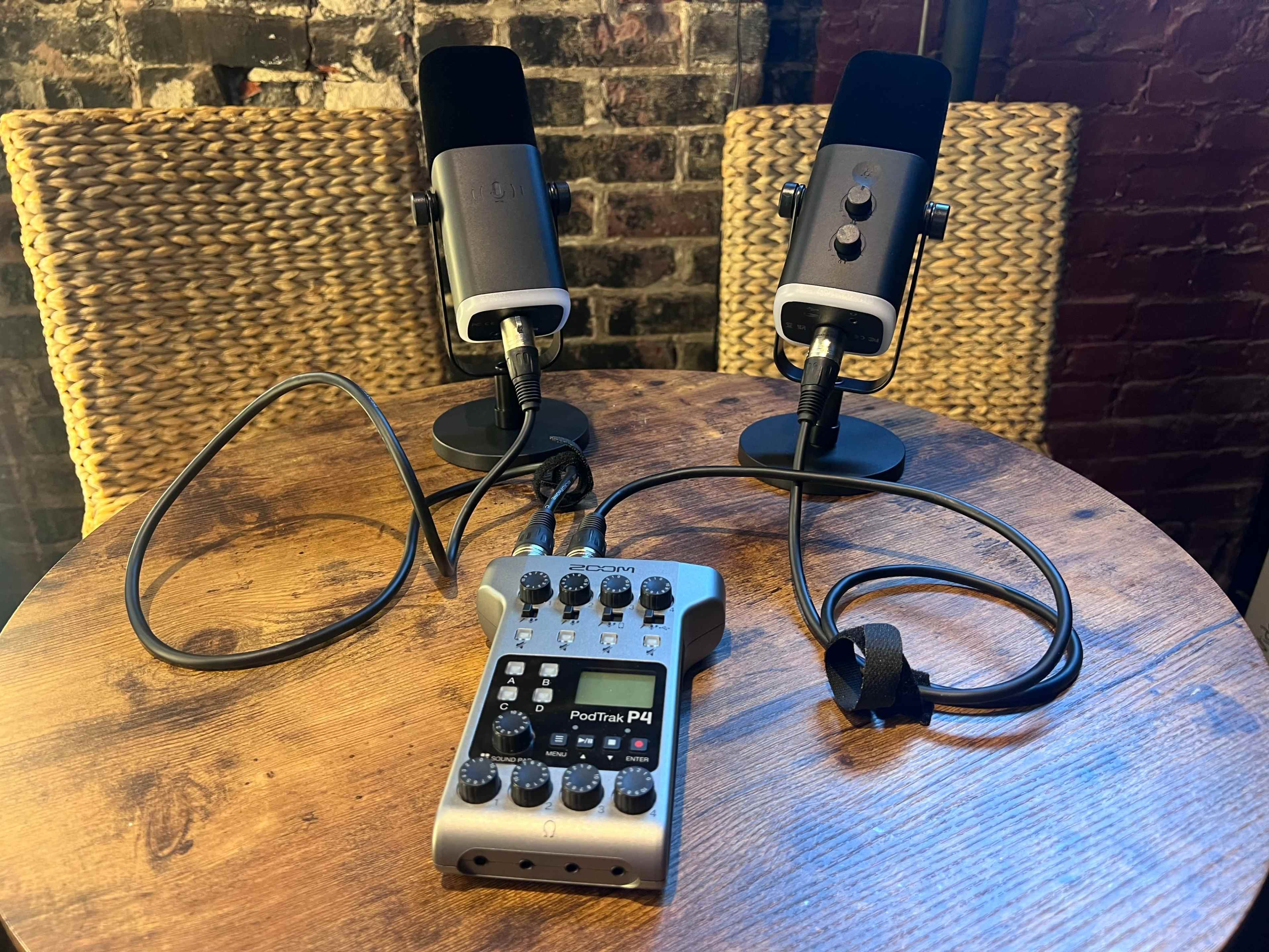 The image shows two microphones mounted on stands connected to a portable audio recorder on a wooden table, with a brick wall in the background.