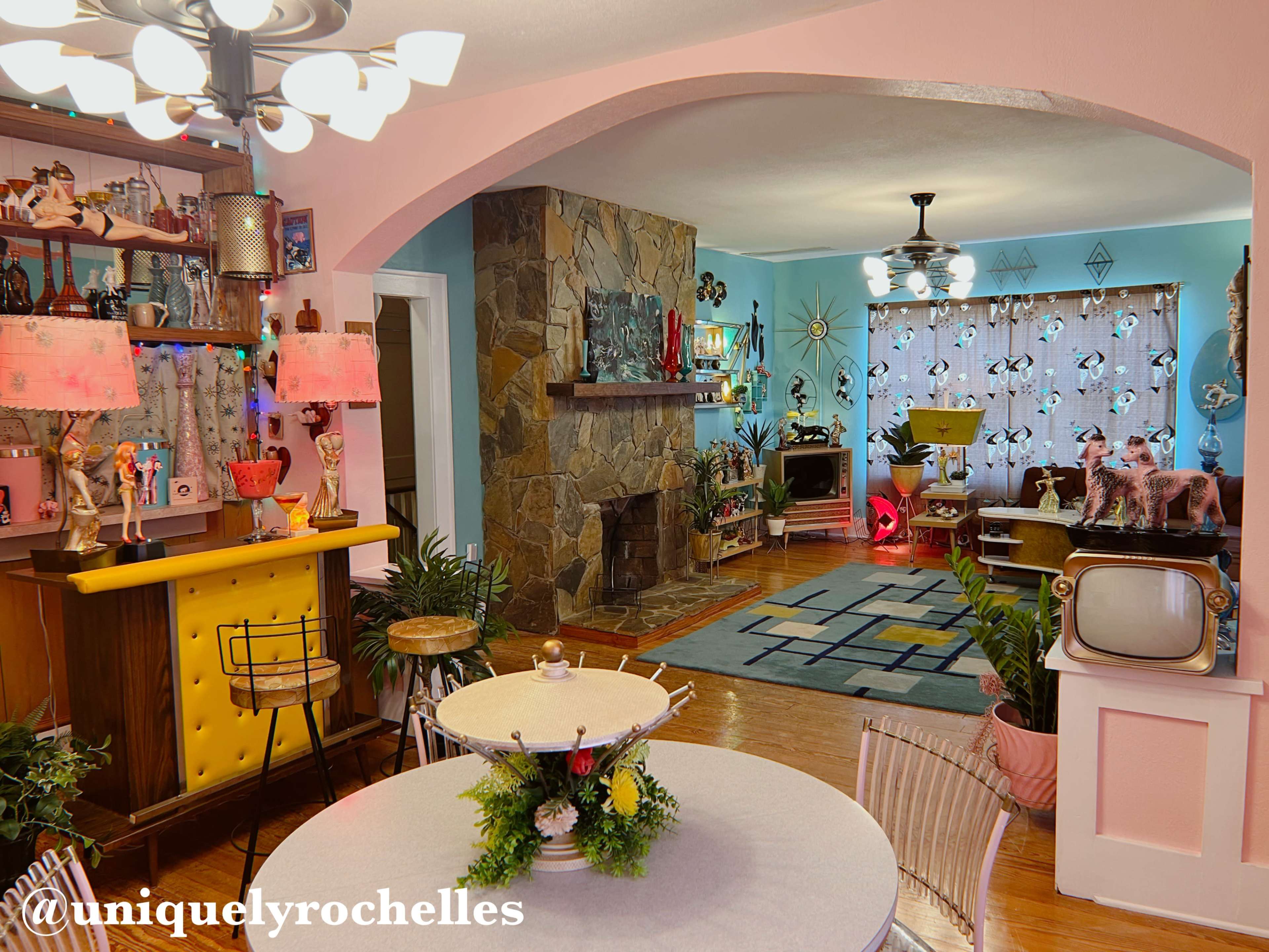 The image showcases a retro-style living space featuring a stone fireplace, colorful decor, and vintage furnishings, including a television and a bar area.