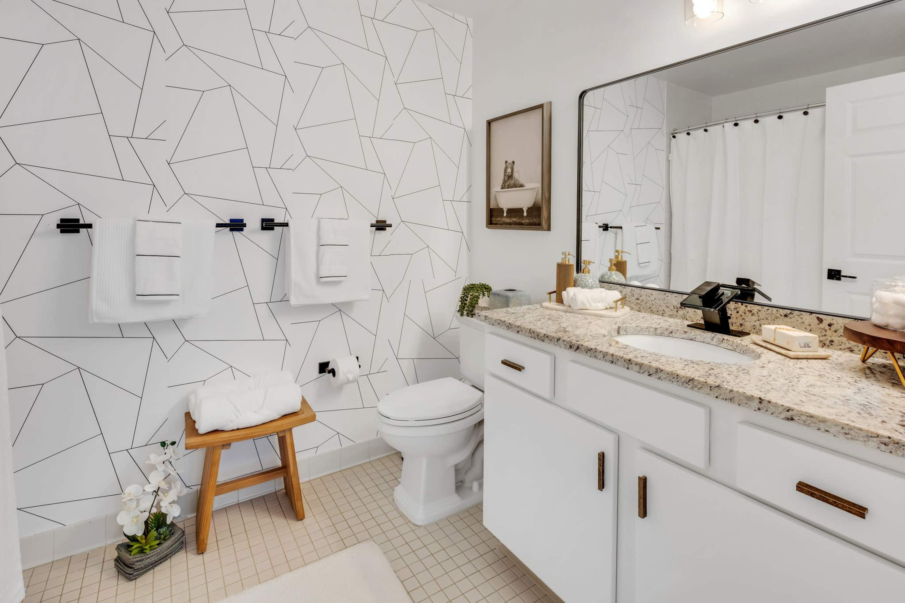The bathroom features a modern design with a geometric patterned wall, a white vanity with a granite countertop, and neatly arranged towels.