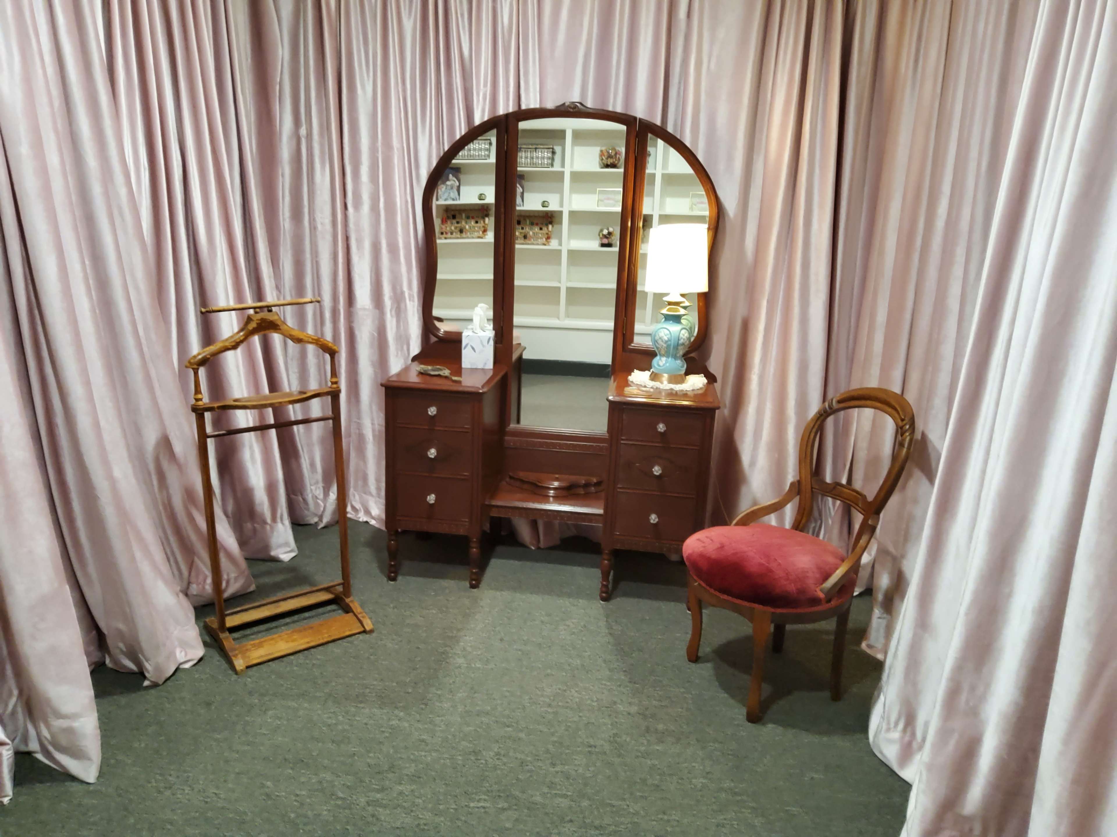 A small room with a dressing table, a mirror, a lamp, a chair, and a clothes rack, all surrounded by light pink curtains.
